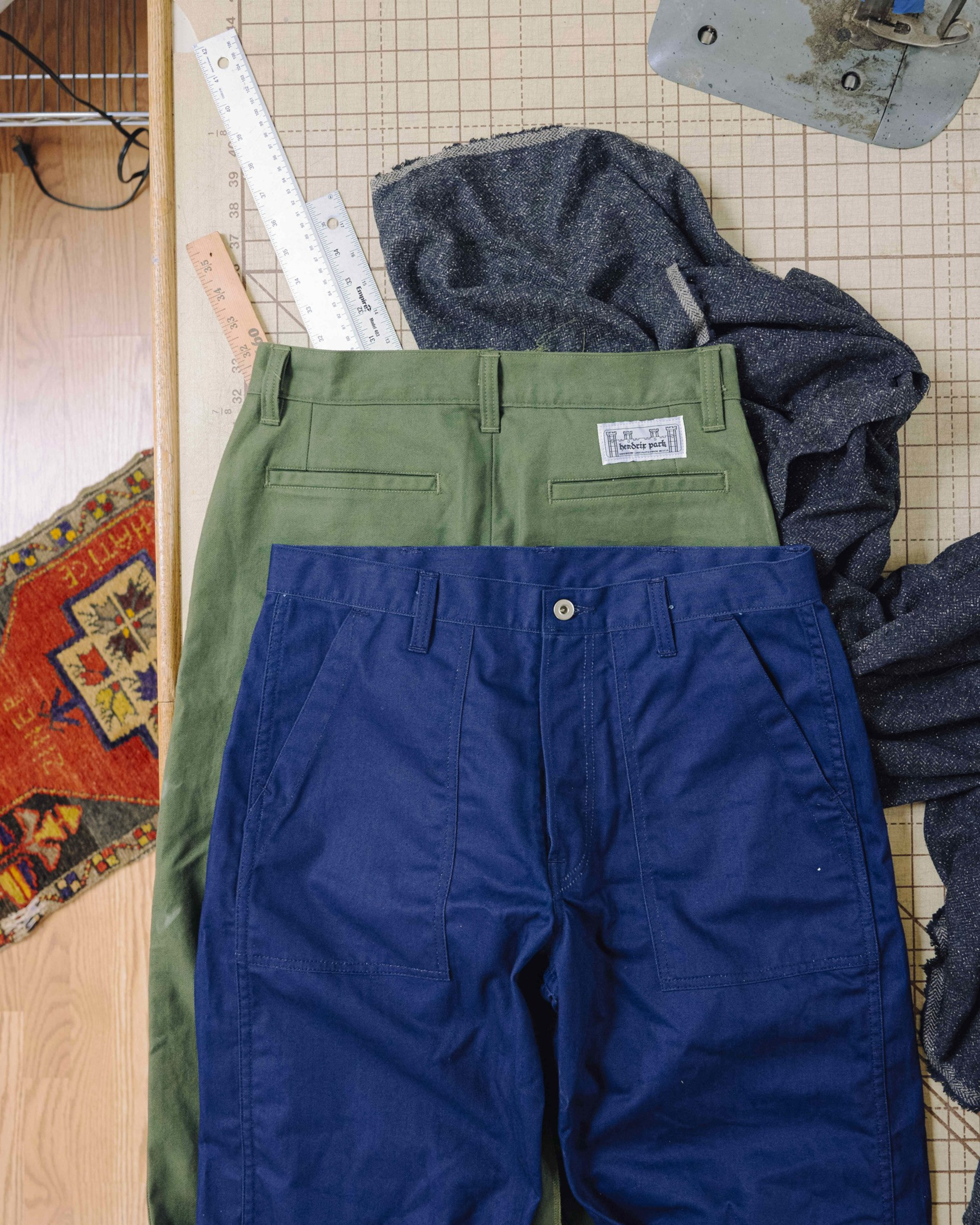 Two pairs of pants, one olive green with a label and one navy blue, lie on a grid-patterned work surface with rulers, fabric, and part of a sewing machine visible.