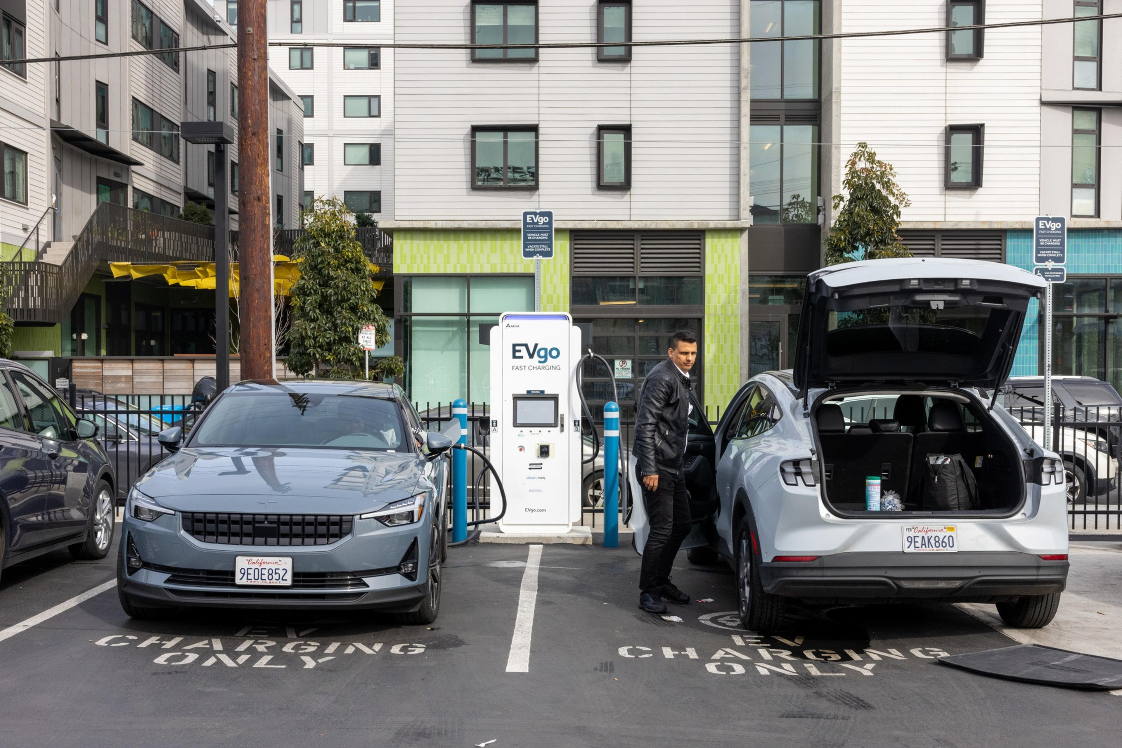 A man stands by two EVs charging at EVgo stations, with "CHARGING ONLY" painted on the ground.