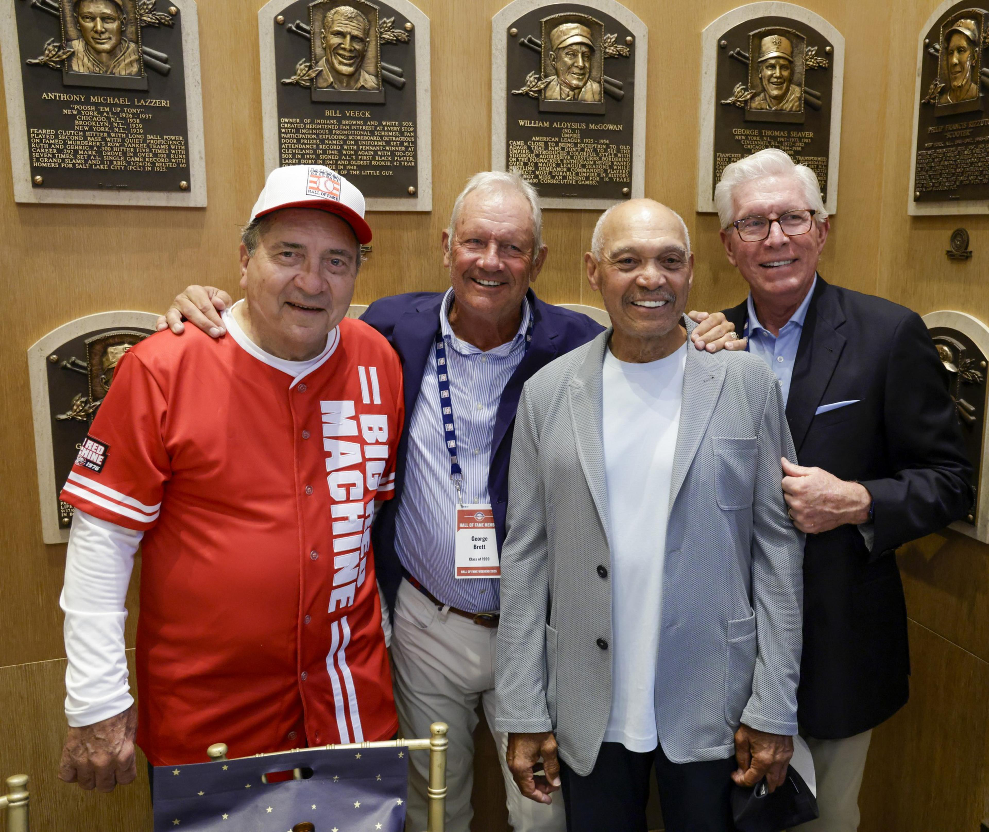 Four men are smiling at the camera, standing in front of a wall displaying plaques. One wears a red &quot;Big Machine&quot; shirt, and another has a &quot;George Brett&quot; nametag.