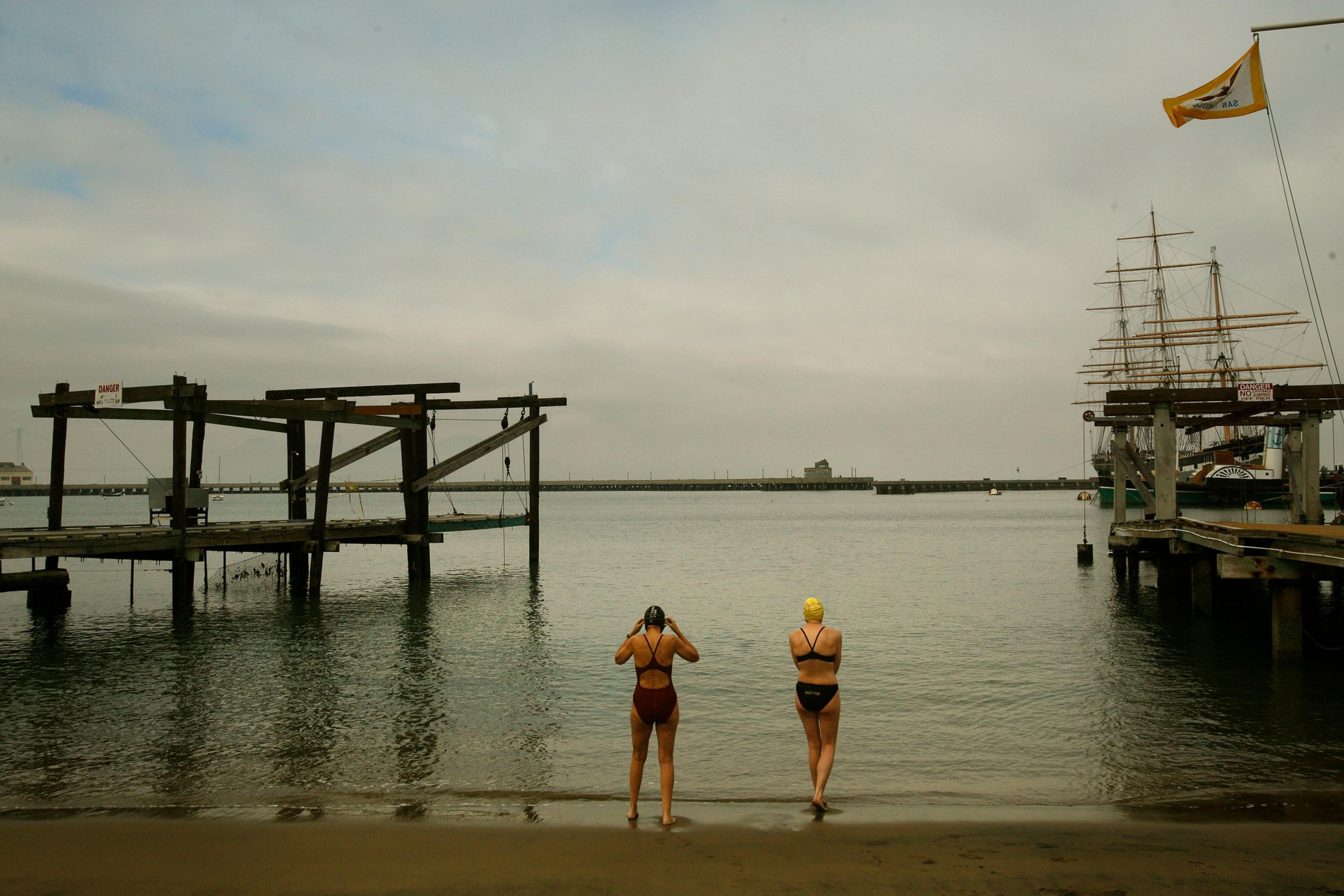 Two swimmers in caps stand at the water’s edge near wooden docks, with a large sailing ship moored on the right under a cloudy sky.