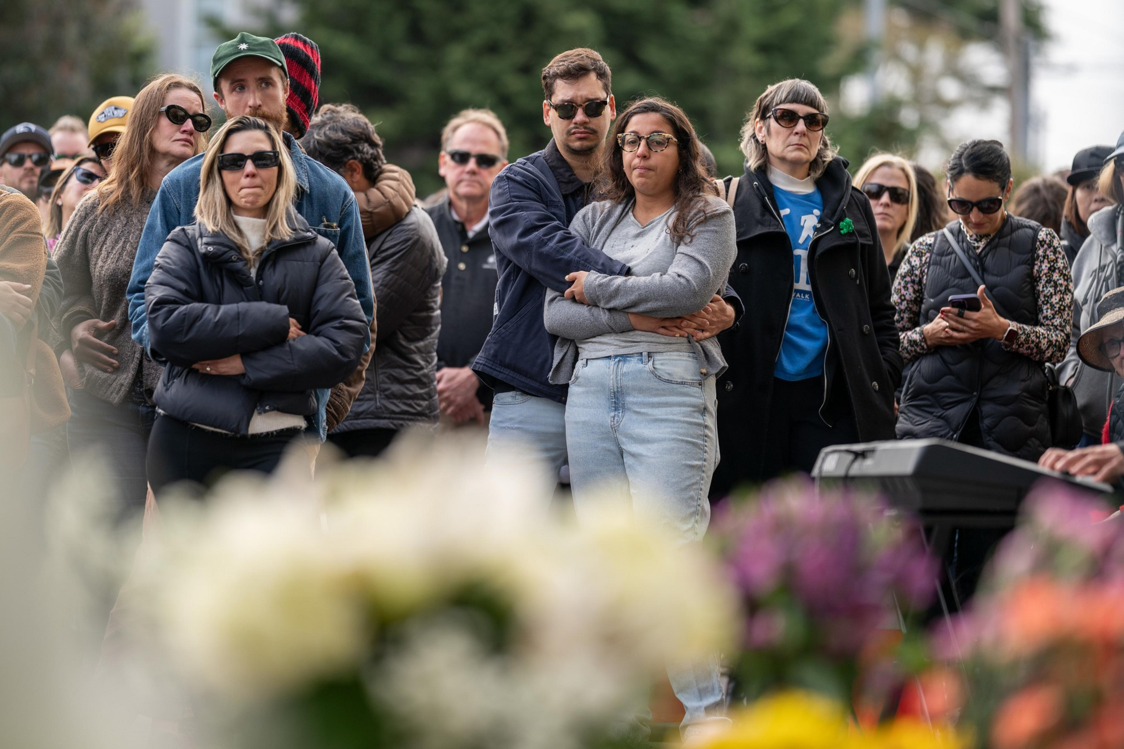 A group of people stand solemnly, many wearing dark clothing and sunglasses. Two individuals are embracing, and flowers in the foreground are slightly out of focus.