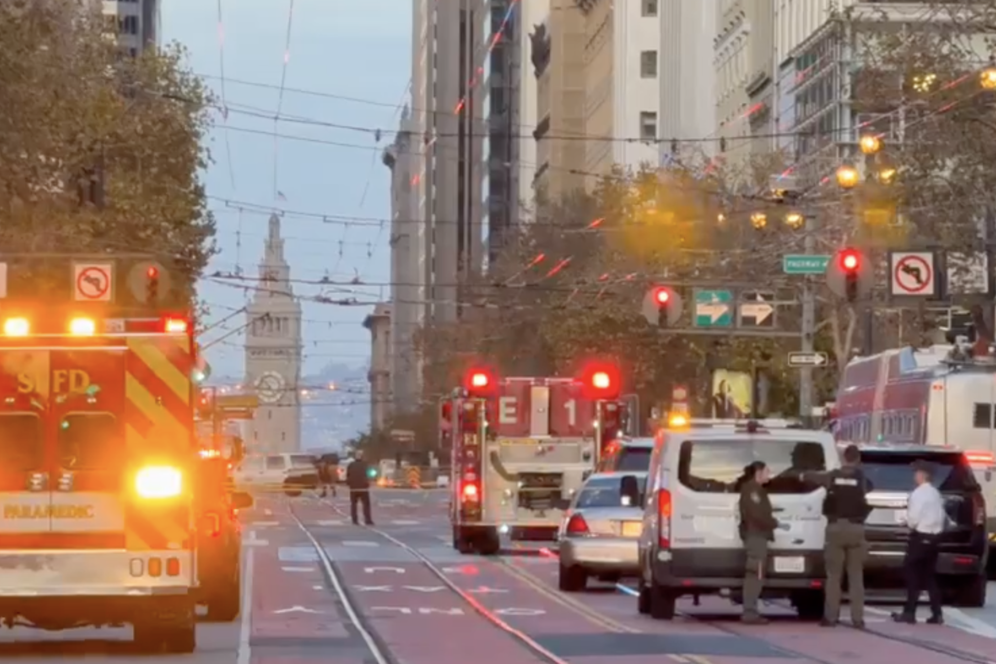 Emergency vehicles with flashing lights block a city street as three people stand near a white van, with a clock tower visible in the distance.