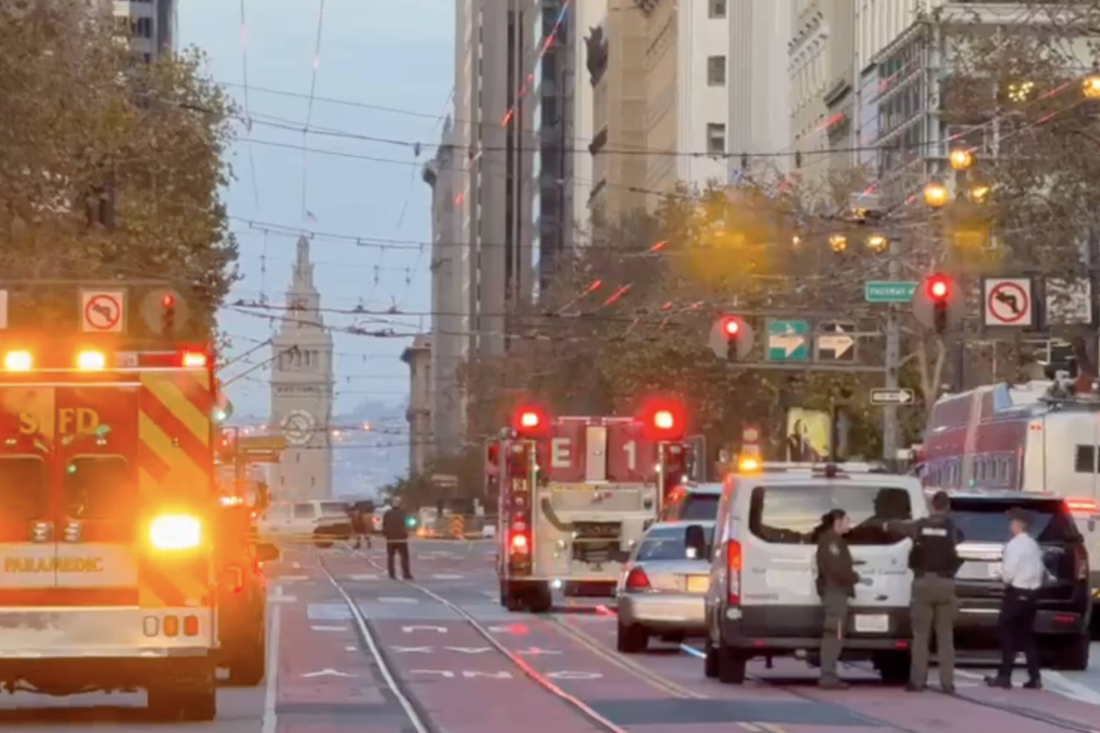 Emergency vehicles with flashing lights block a city street as three people stand near a white van, with a clock tower visible in the distance.