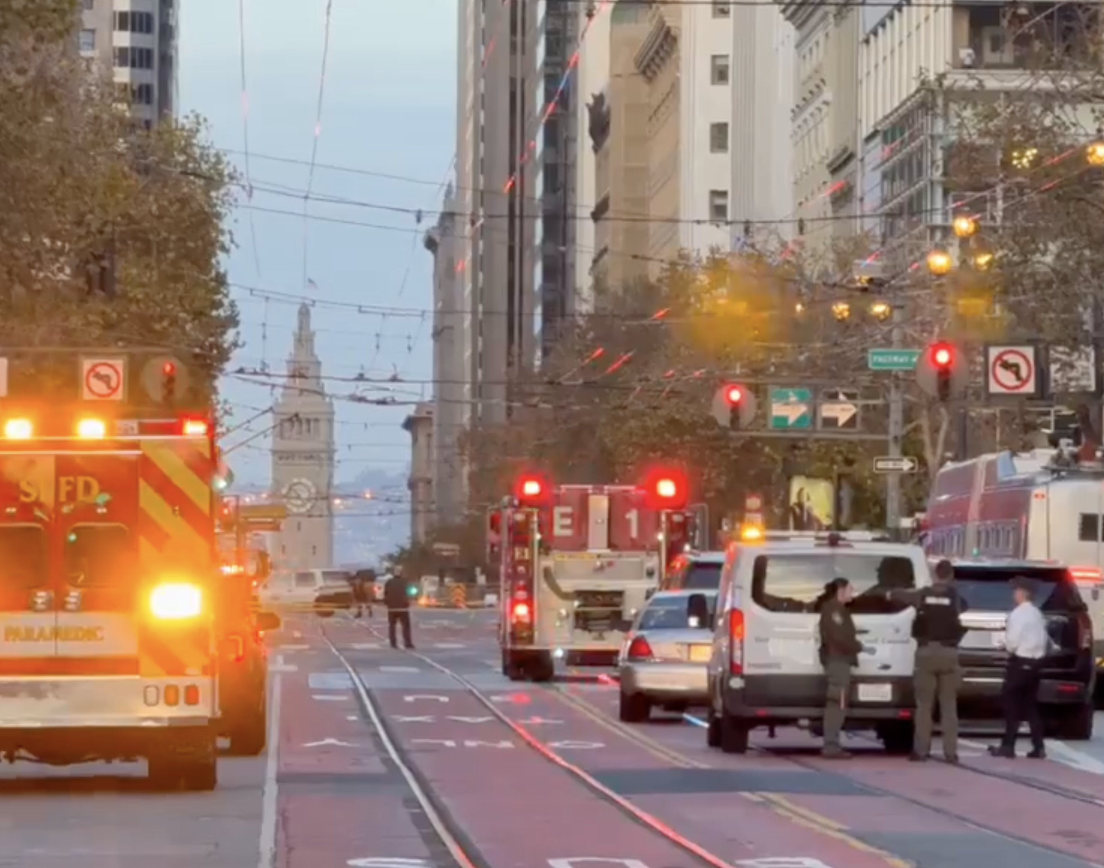 Emergency vehicles with flashing lights block a city street as three people stand near a white van, with a clock tower visible in the distance.
