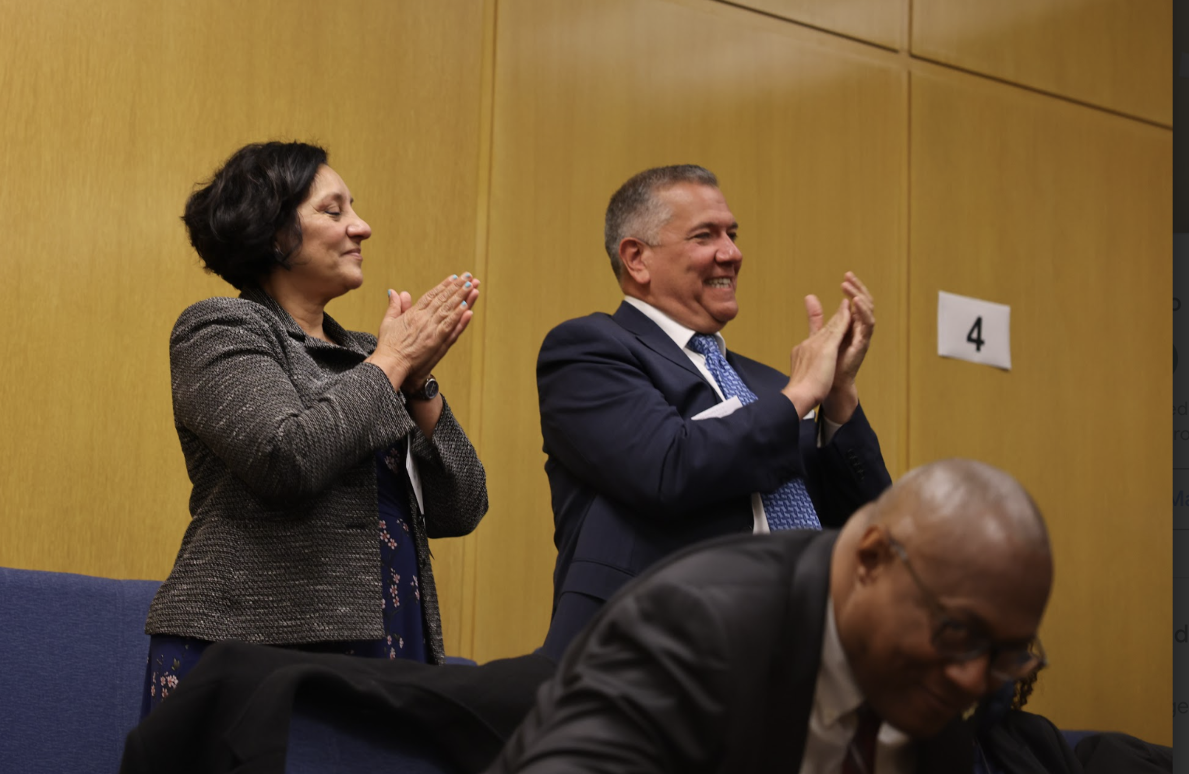 A woman and a man are standing and clapping happily, while another man is seated and leaning forward, all in a room with wooden paneling and a sign with the number 4.