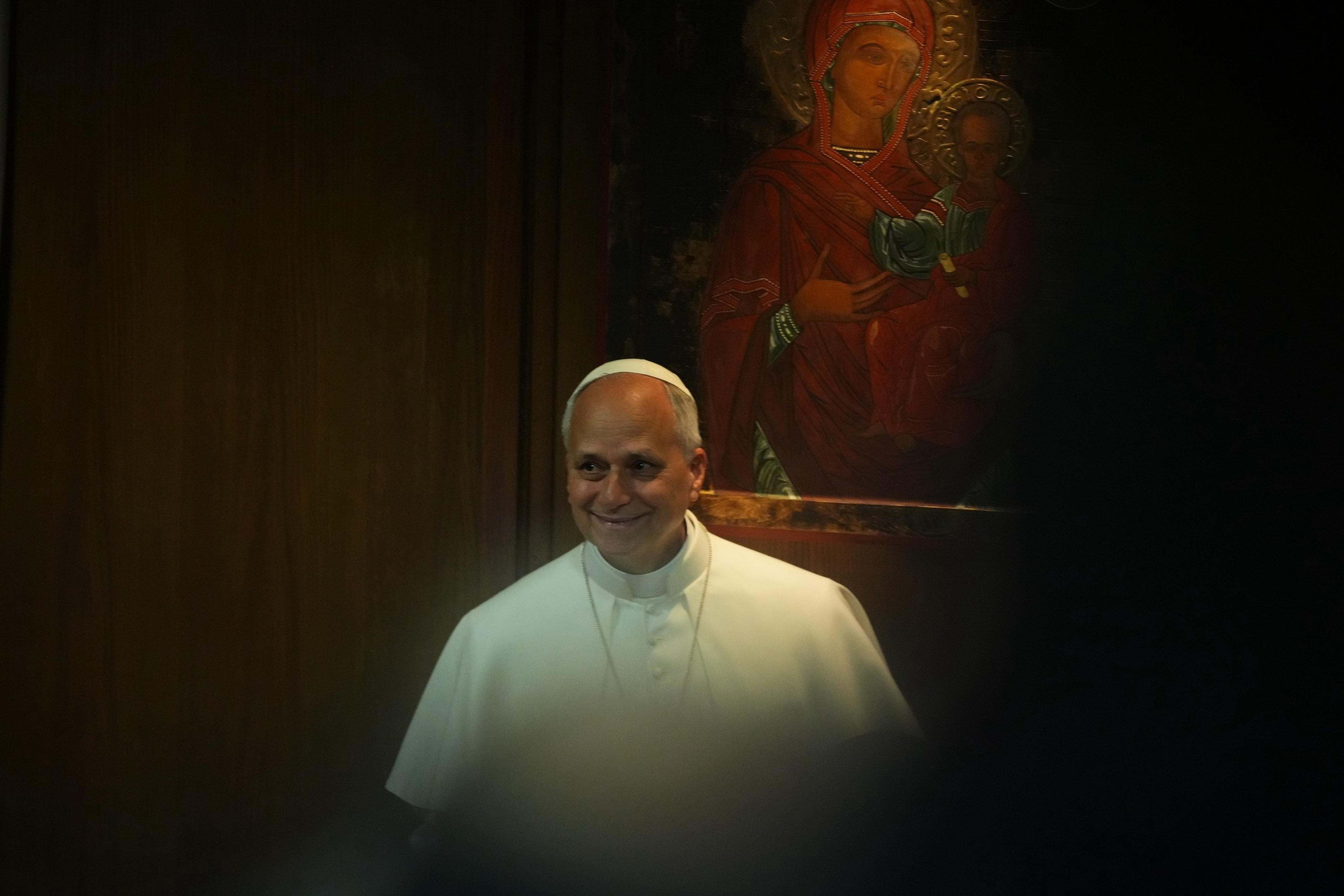 A man in white religious attire with a white skullcap smiles gently, standing near a wooden wall and an icon of a woman holding a child.