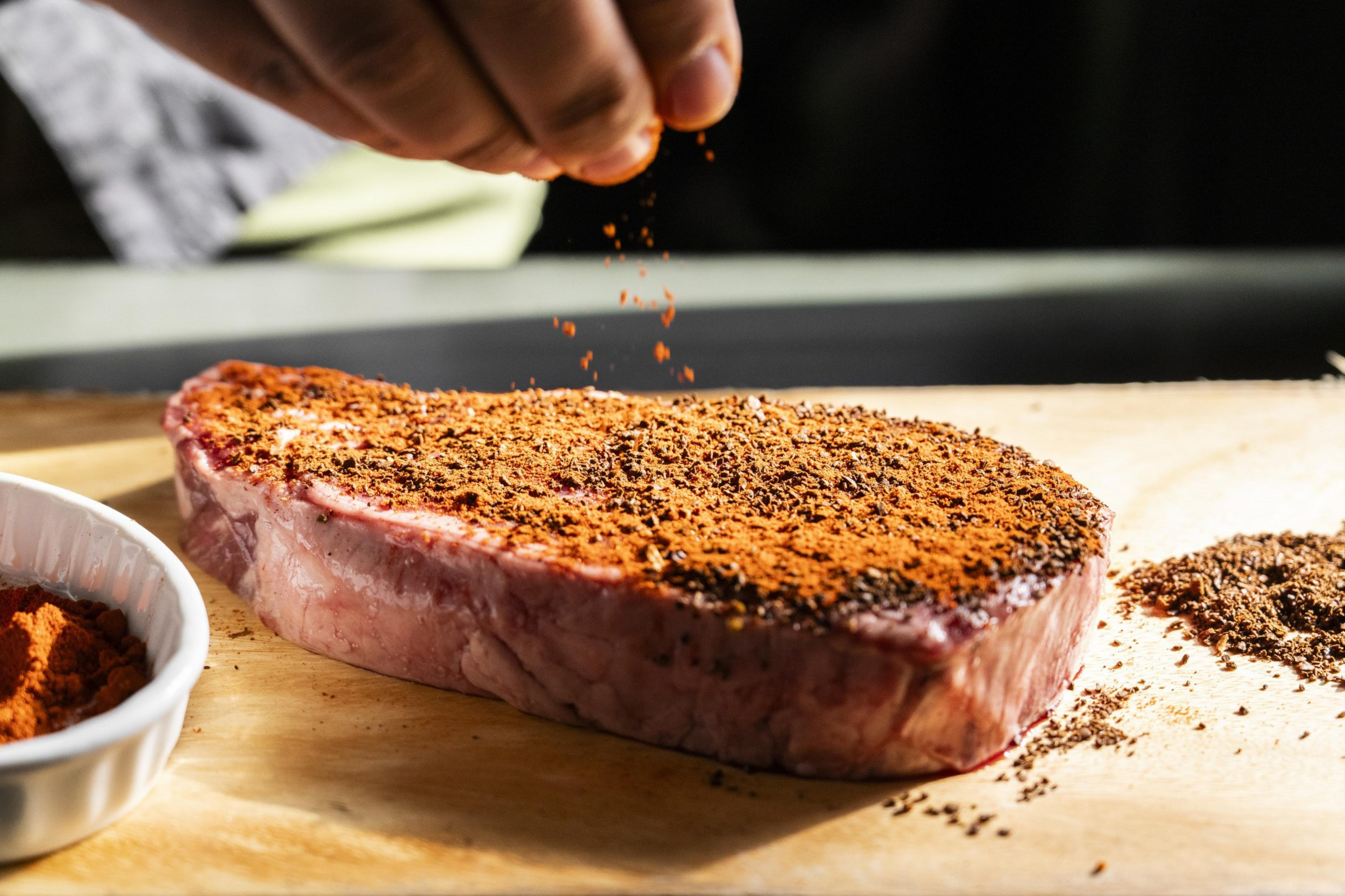 A hand sprinkles seasonings onto a raw steak on a wooden board. The steak is coated with reddish spices, and a small dish of similar spices is nearby.