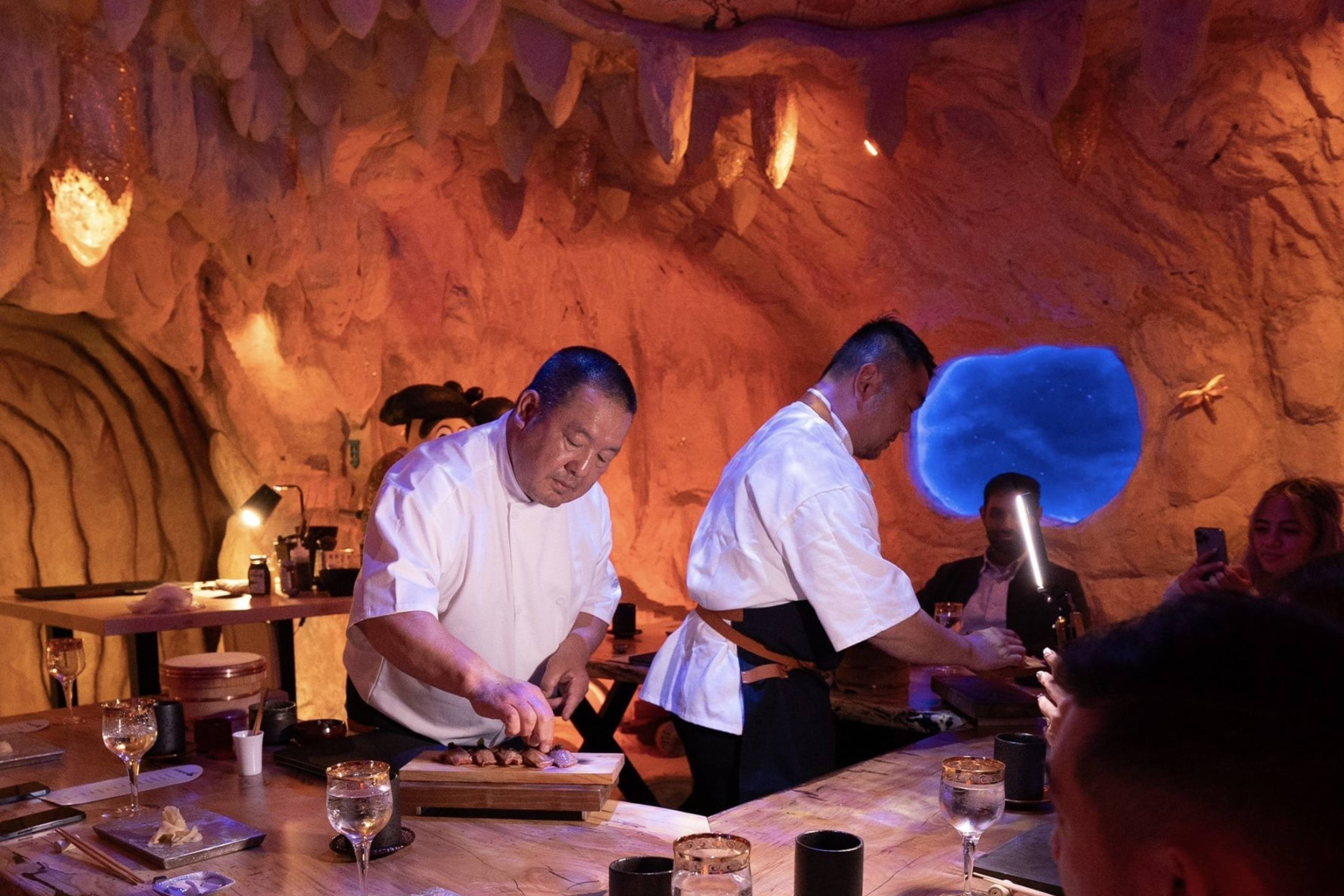 Two chefs in white attire prepare food on a wooden table, surrounded by diners in a cave-themed restaurant with warm lighting and wall decorations.