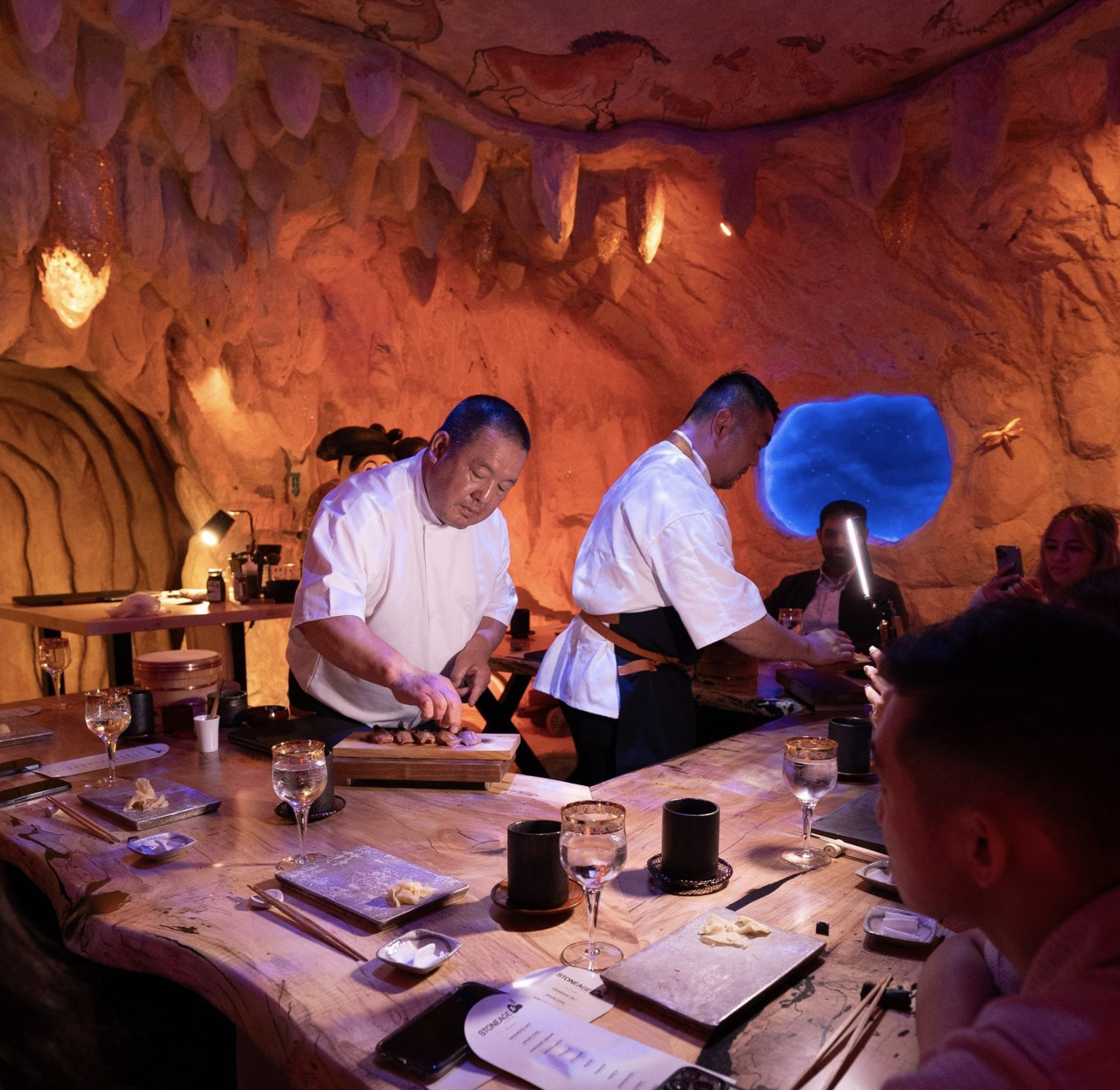 Two chefs in white attire prepare food on a wooden table, surrounded by diners in a cave-themed restaurant with warm lighting and wall decorations.