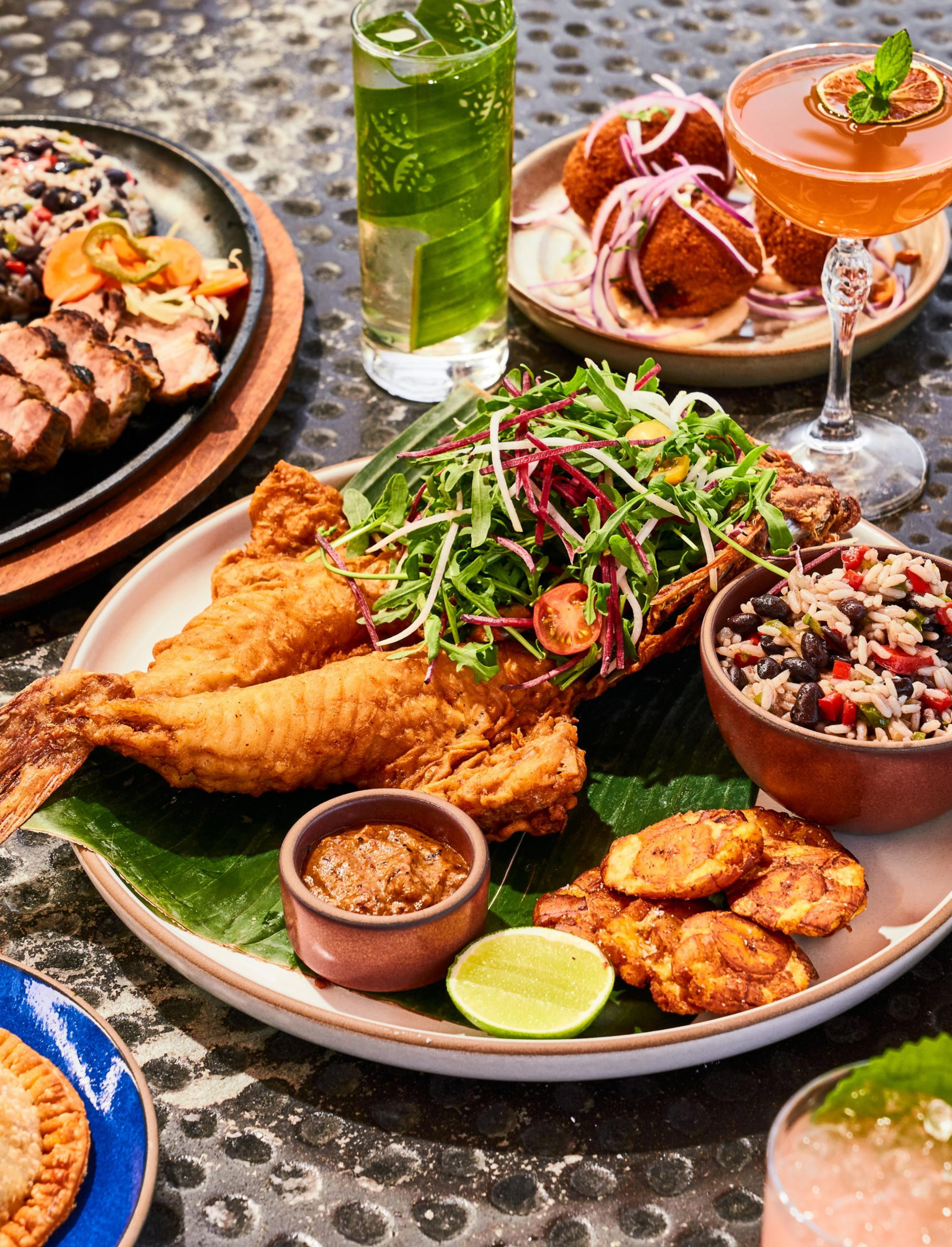 A plate with fried fish topped with greens, rice, plantains, sauce, and lime slices. Nearby are drinks, meat, and fried balls with onions.