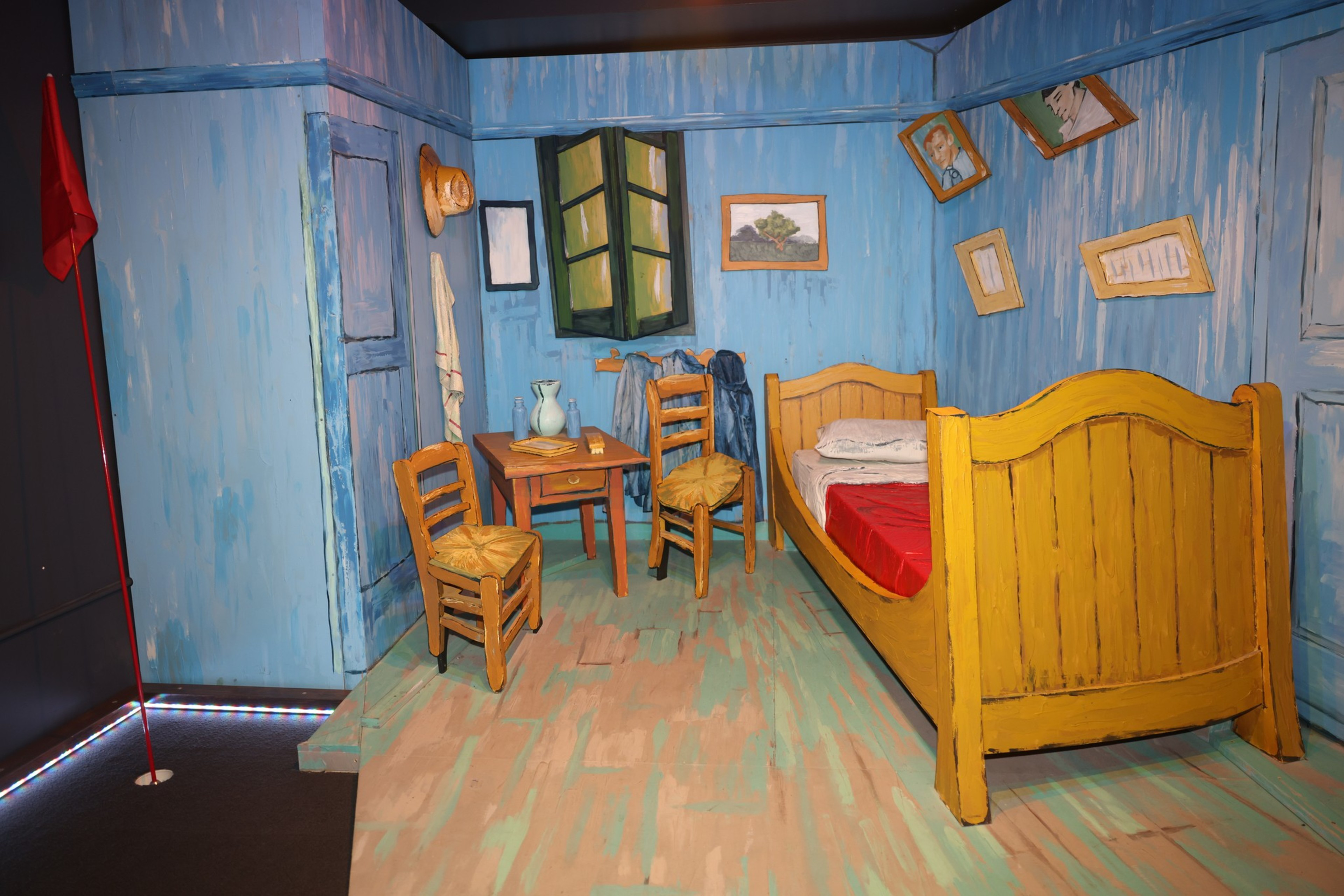 The image depicts a 3D recreation of Van Gogh's painting &quot;Bedroom in Arles,&quot; featuring blue walls, a wooden bed, chairs, a table, green window, hat, and tilted wall frames.