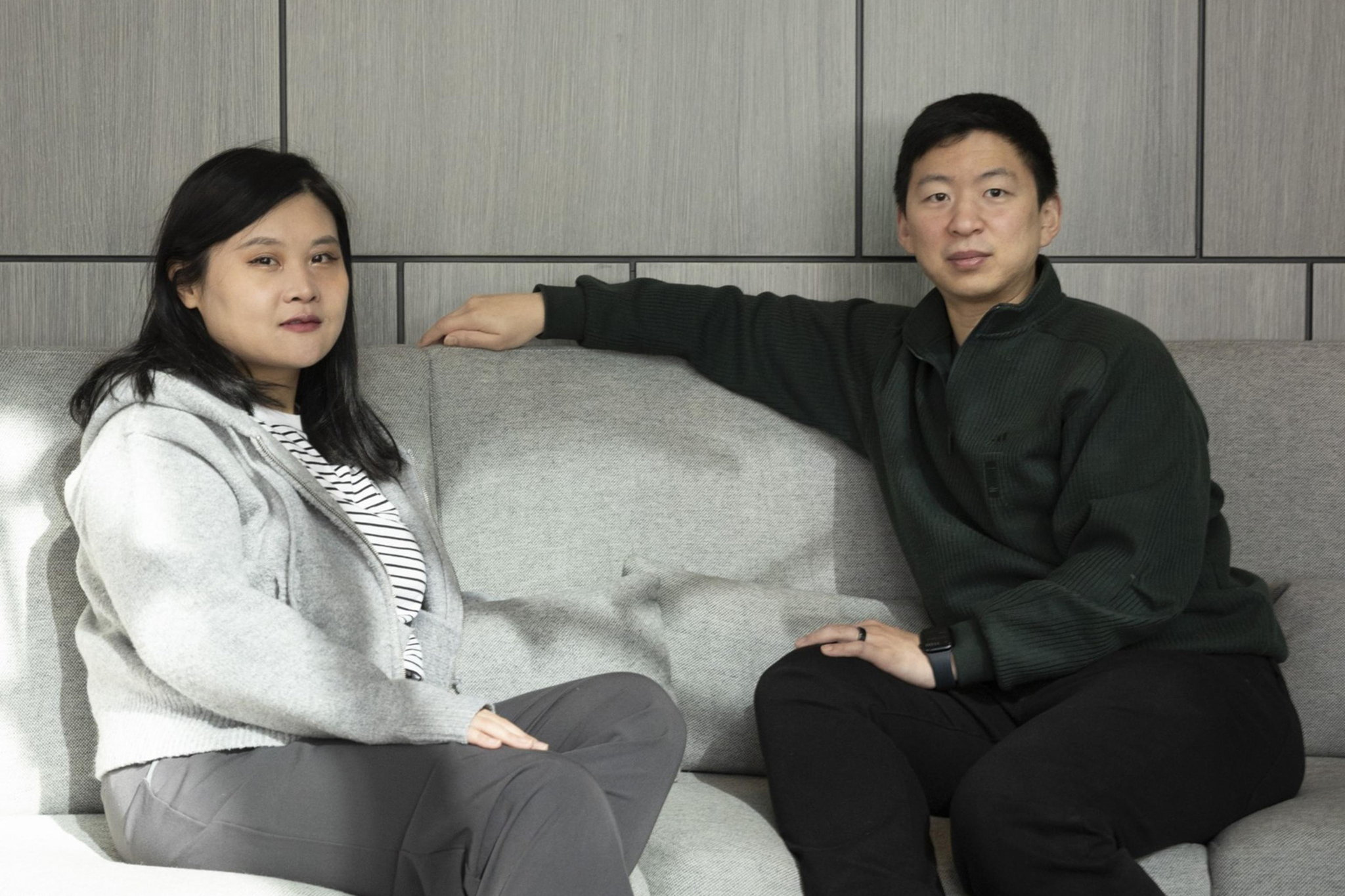 Two people sit on a gray sofa in a modern room with paneled walls. One wears a gray sweater and striped shirt, the other a dark sweater and pants. Both wear sneakers.