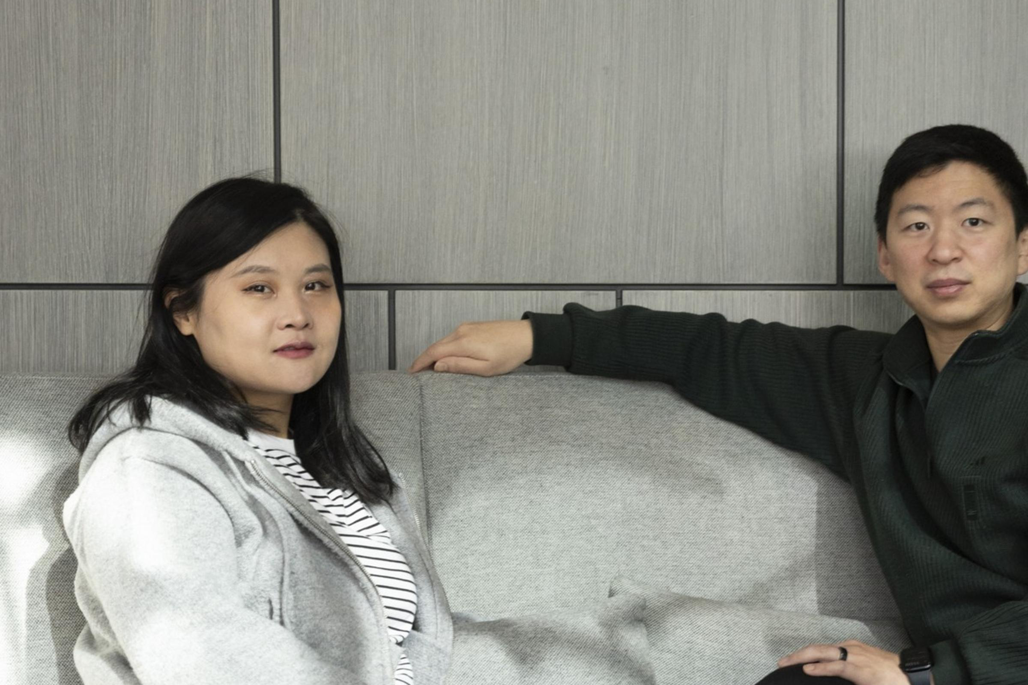 Two people sit on a gray sofa in a modern room with paneled walls. One wears a gray sweater and striped shirt, the other a dark sweater and pants. Both wear sneakers.
