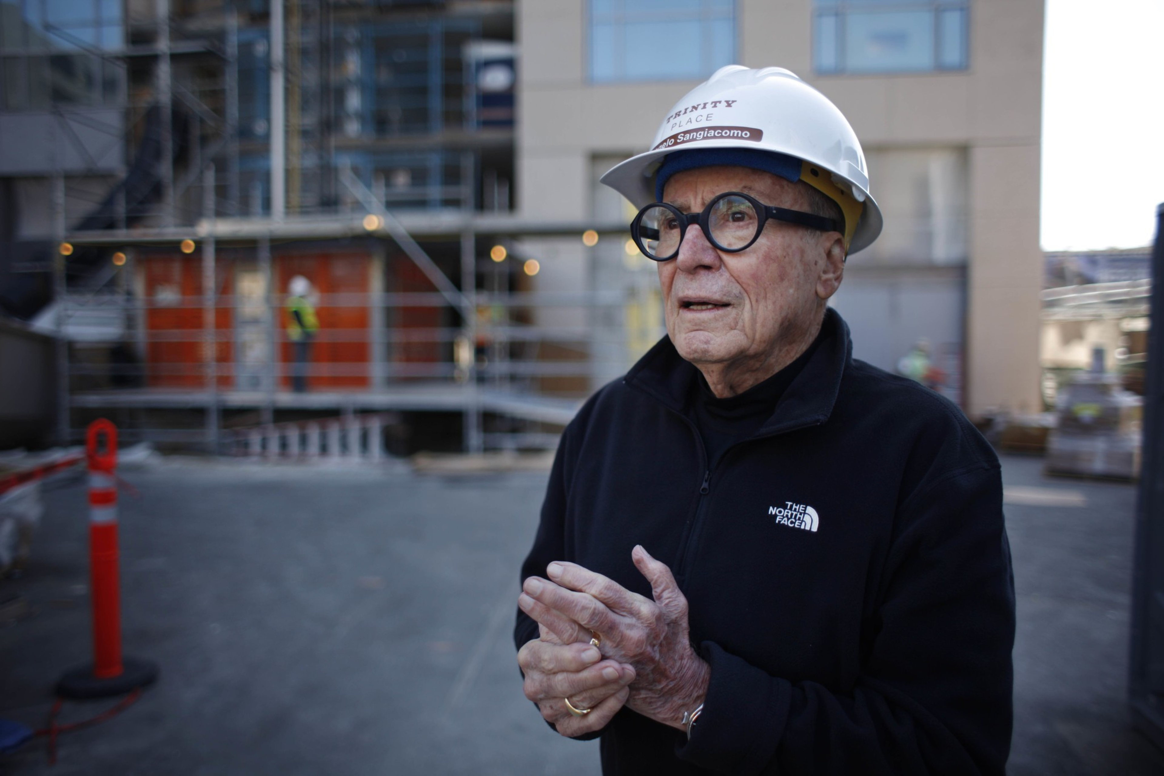 An elderly man wearing a white hard hat and black North Face jacket stands at a construction site, with scaffolding and workers blurred in the background.