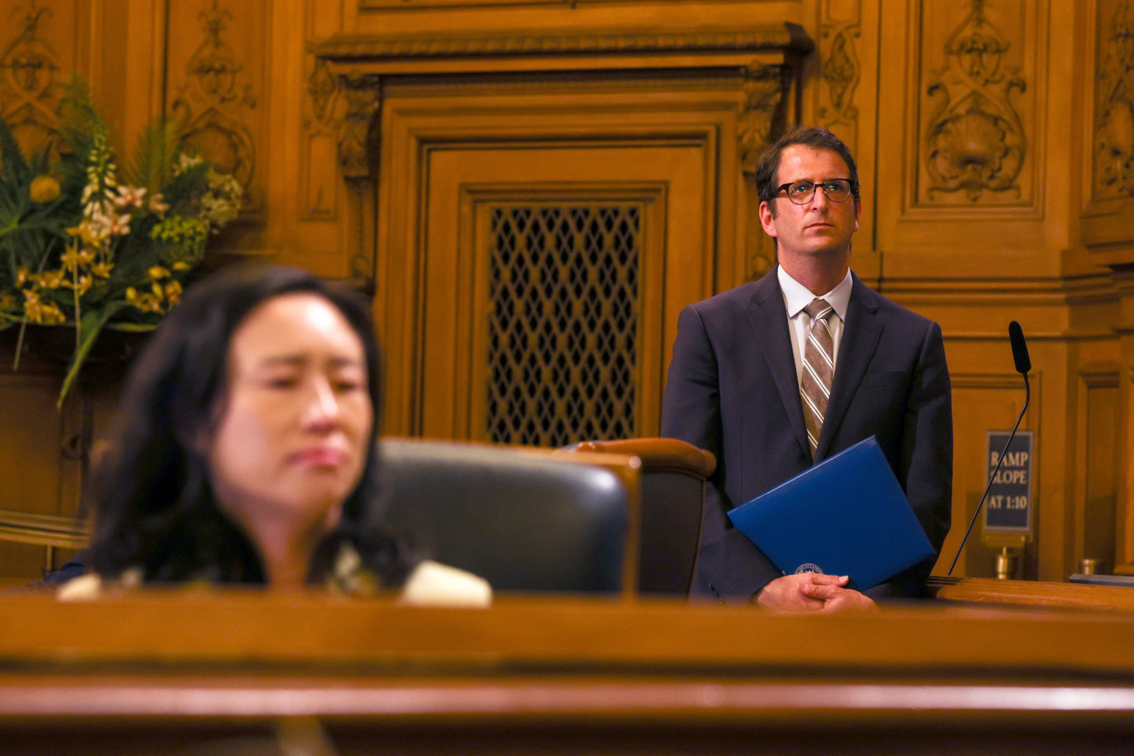 A man in a suit with glasses holds a blue folder standing behind a woman seated in a courtroom with wooden paneling.