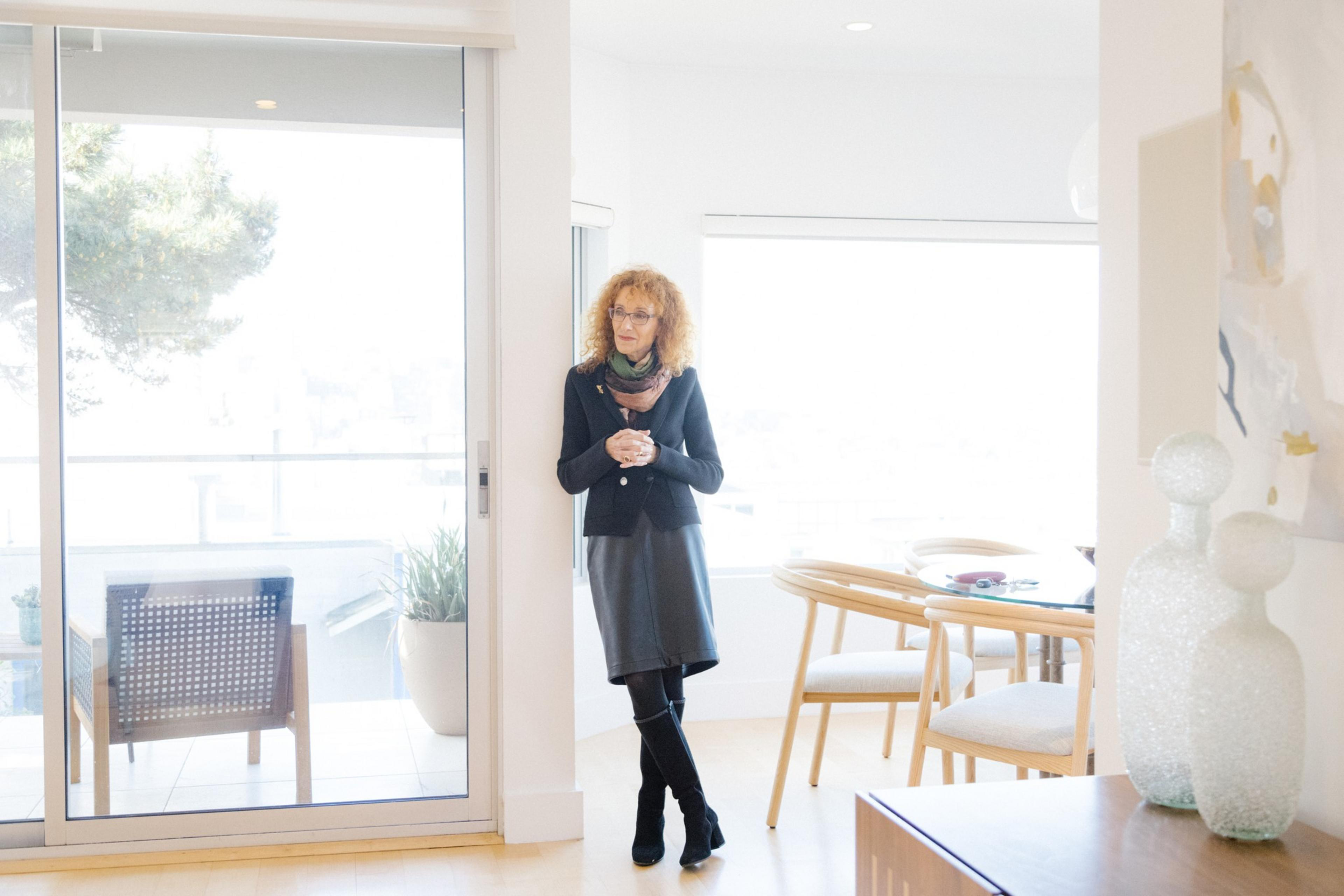 A woman stands in a bright room near an open door, with outdoor seating visible outside and modern decor inside.