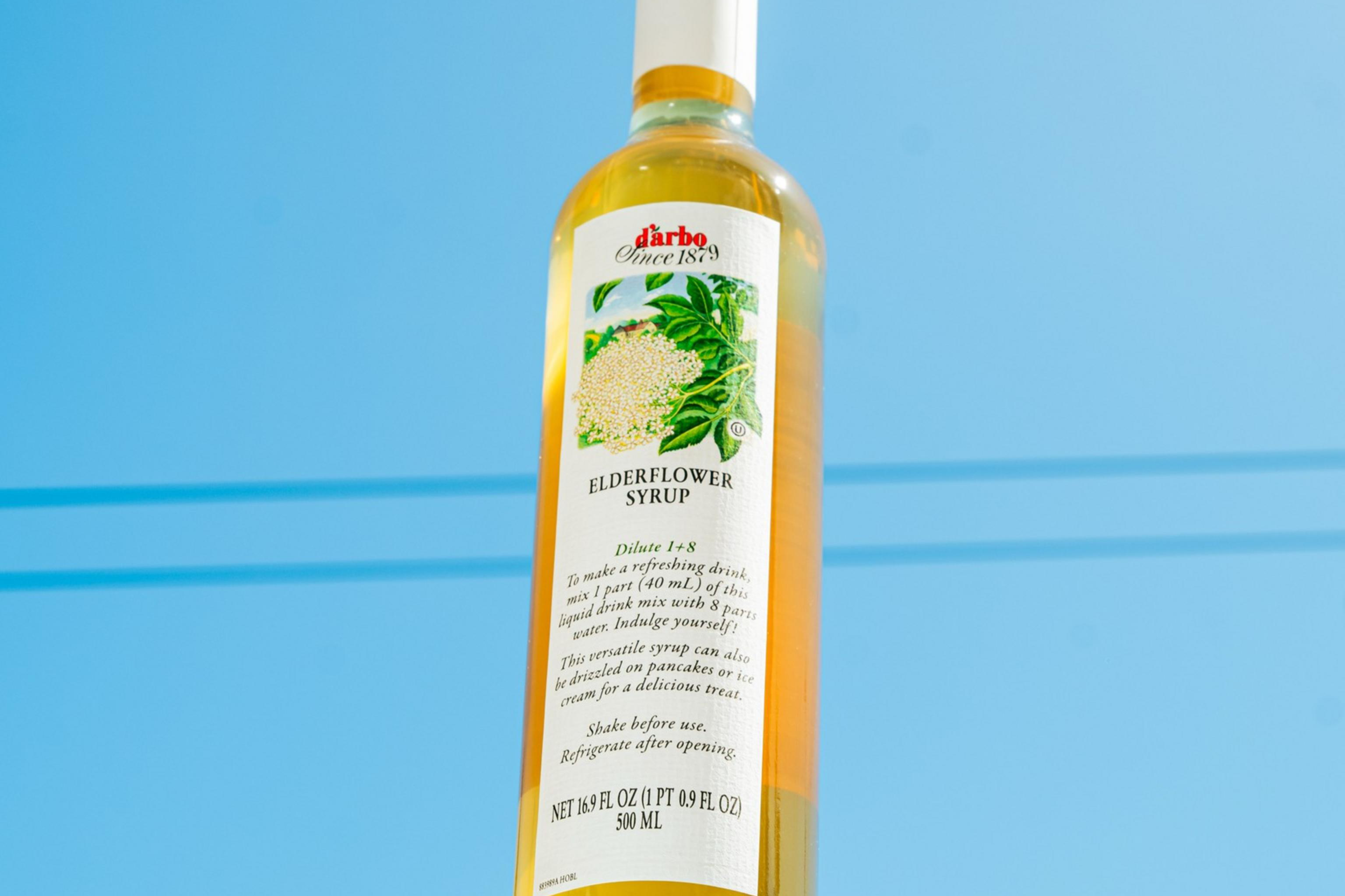 A bottle of elderflower syrup with a white cap sits against a clear blue sky. The label features illustrations of elderflowers and green leaves.