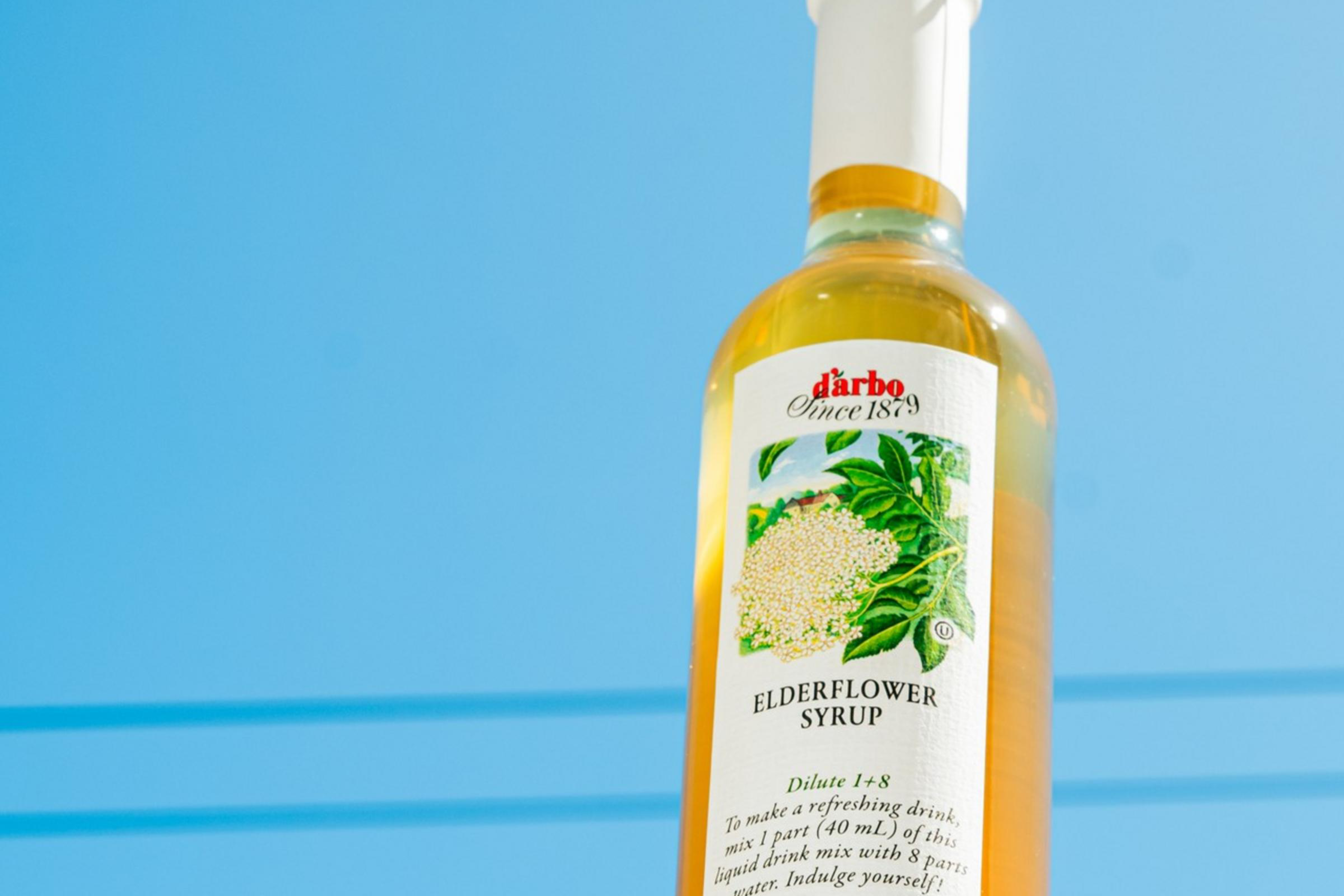 A bottle of elderflower syrup with a white cap sits against a clear blue sky. The label features illustrations of elderflowers and green leaves.