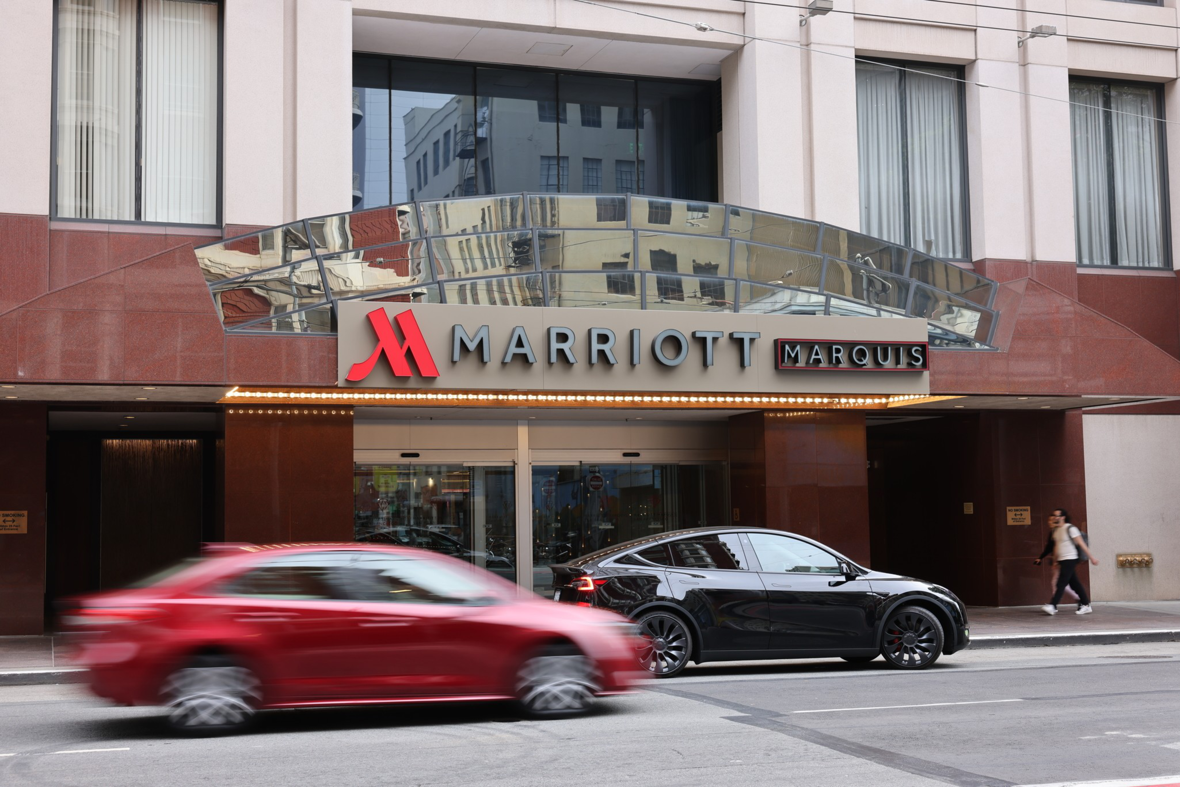 The image shows the front of the Marriott Marquis hotel, with its sign and entrance. A black car is parked on the street, and a red car is driving by.