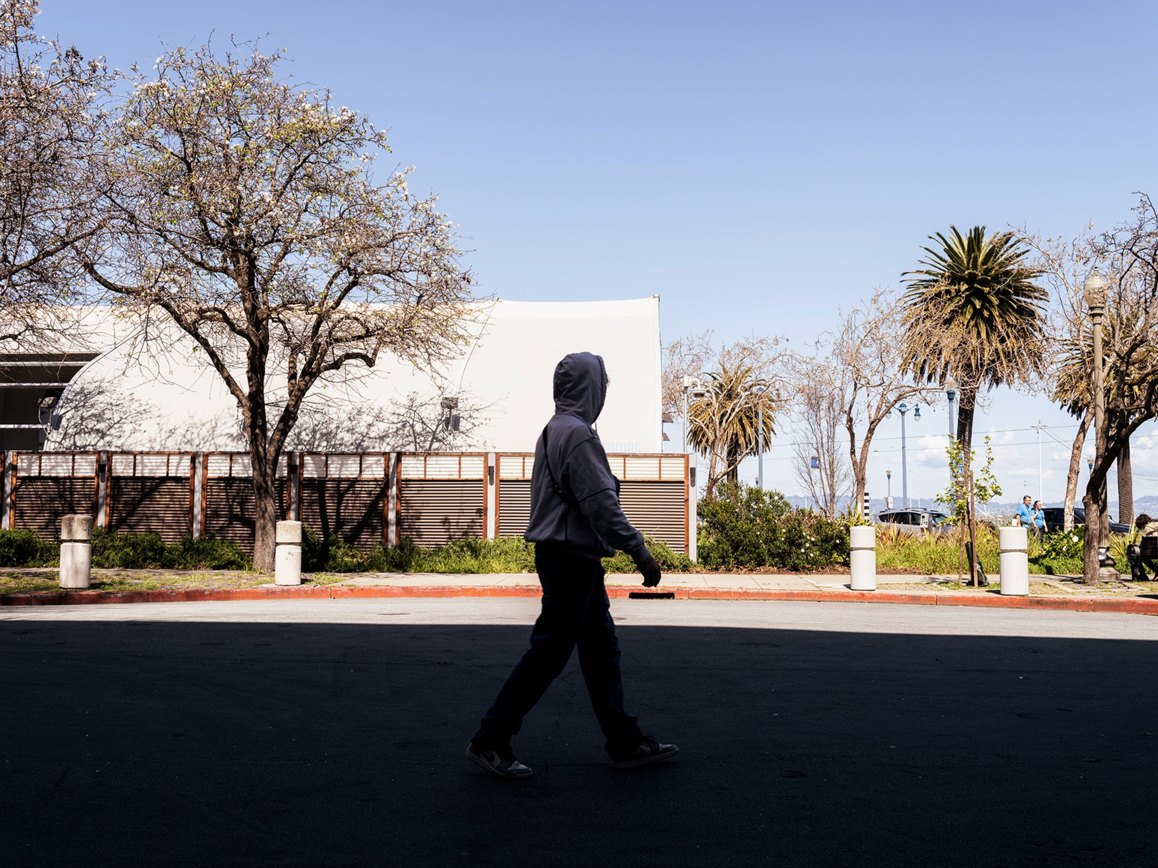 A person in a hoodie walks along a shaded street flanked by leafless trees. The background shows a clear sky and a building.