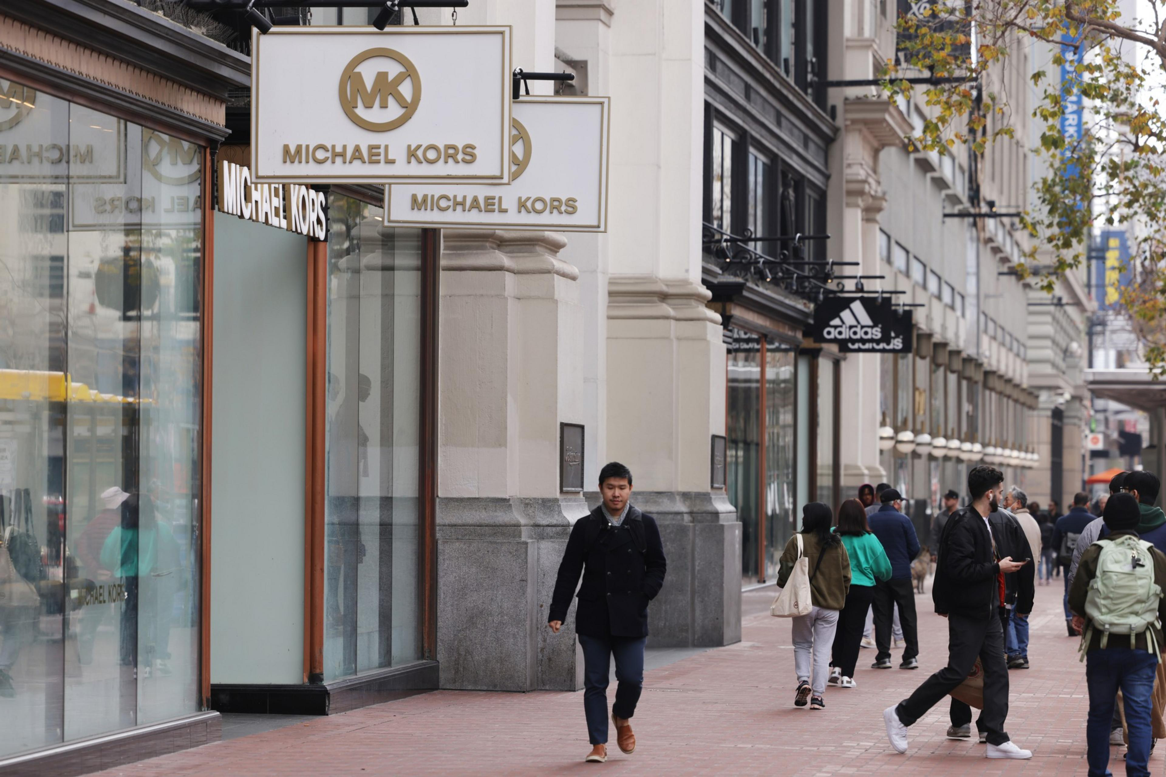 A busy city street with pedestrians and storefronts including Michael Kors and Adidas.