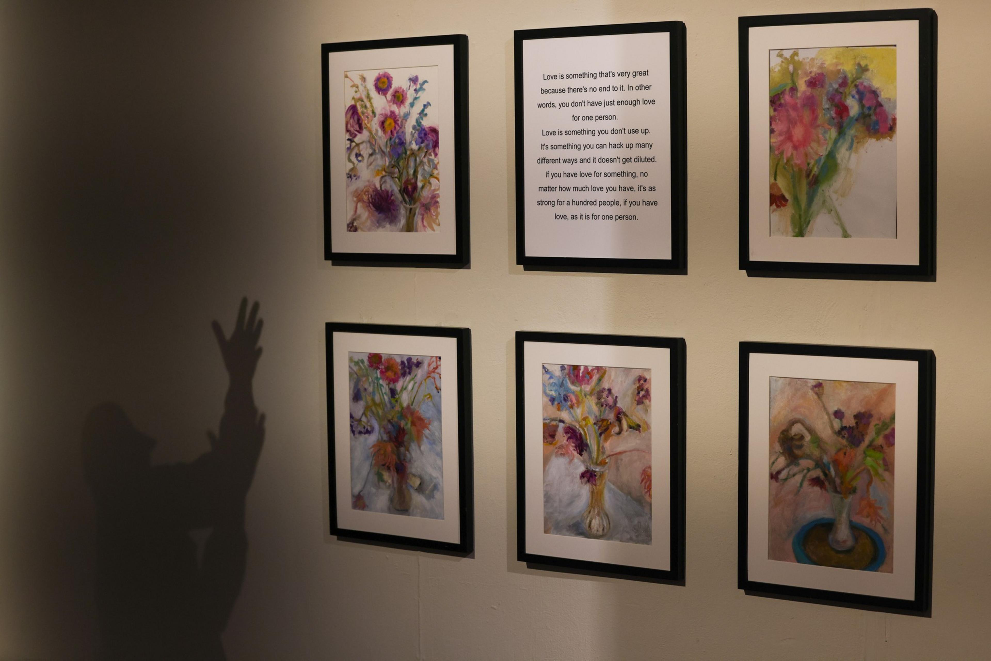 Six framed pictures hang on a wall, five depicting colorful flower bouquets and one with a poem about love, while a shadow of a person raising their hand appears on the left.