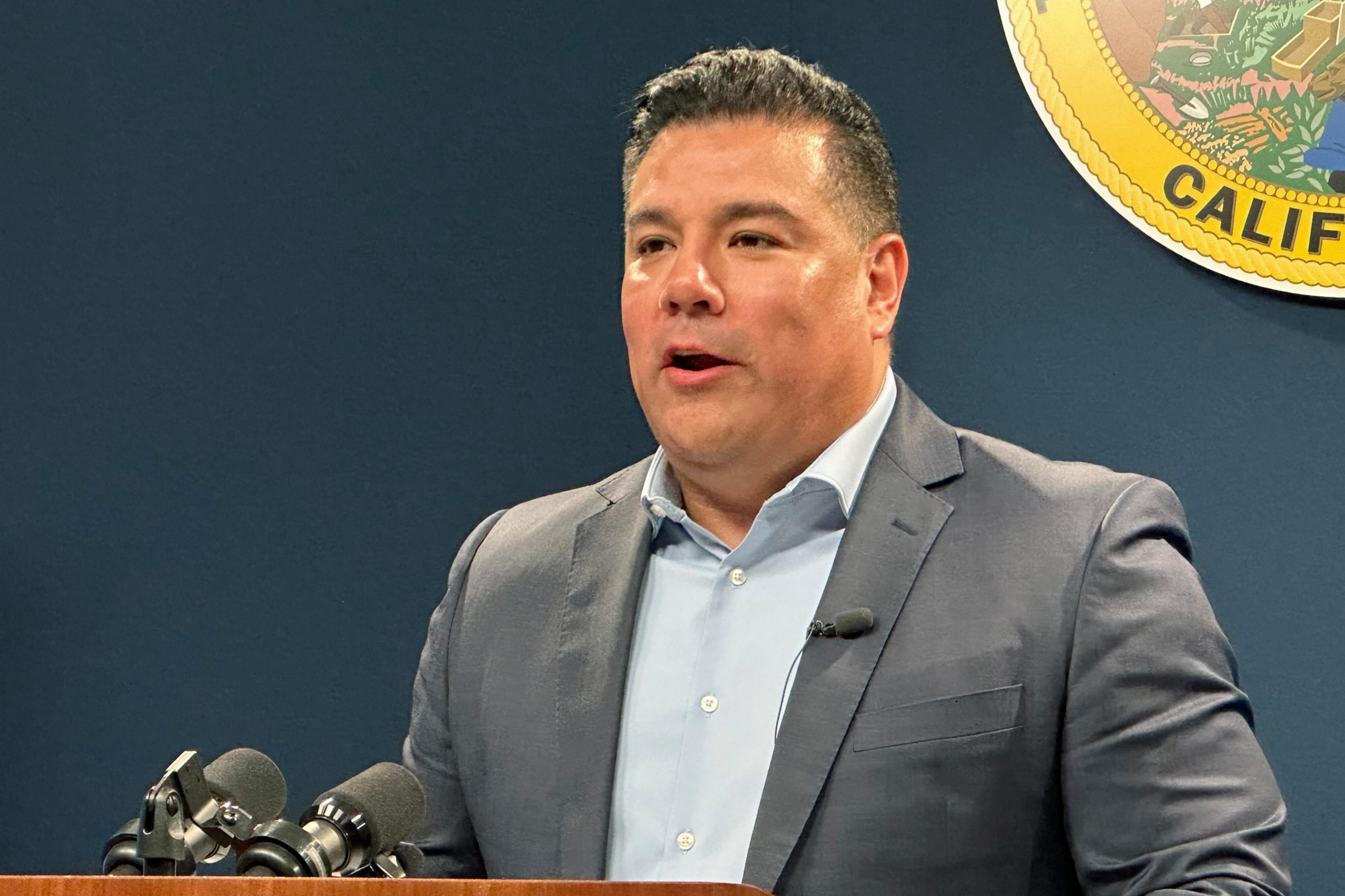 Man in blue collared shirt and gray jacket speaks into microphone