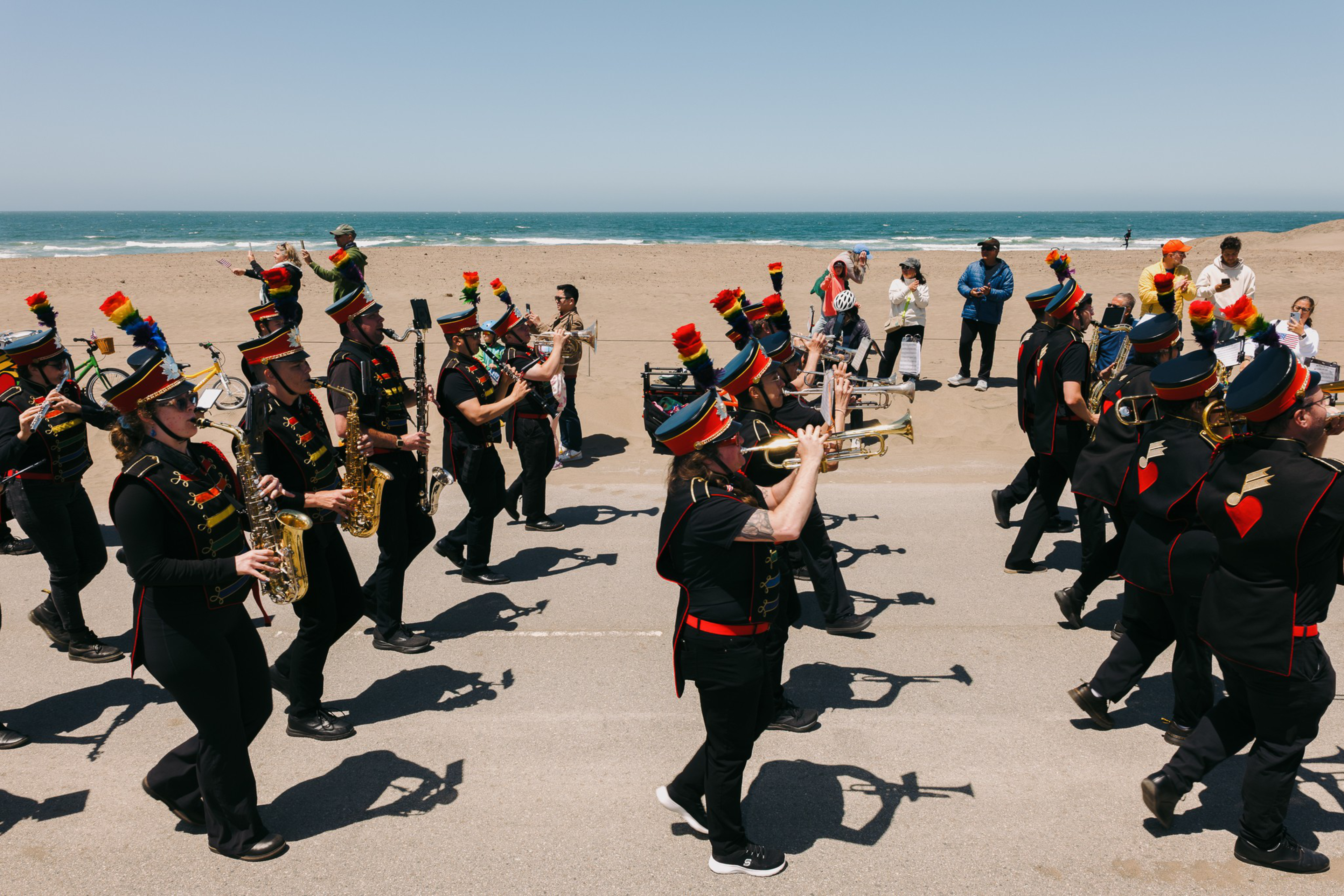 A marching band in colorful uniforms plays instruments on a beachside path, as people watch and the ocean stretches out in the background under a clear sky.