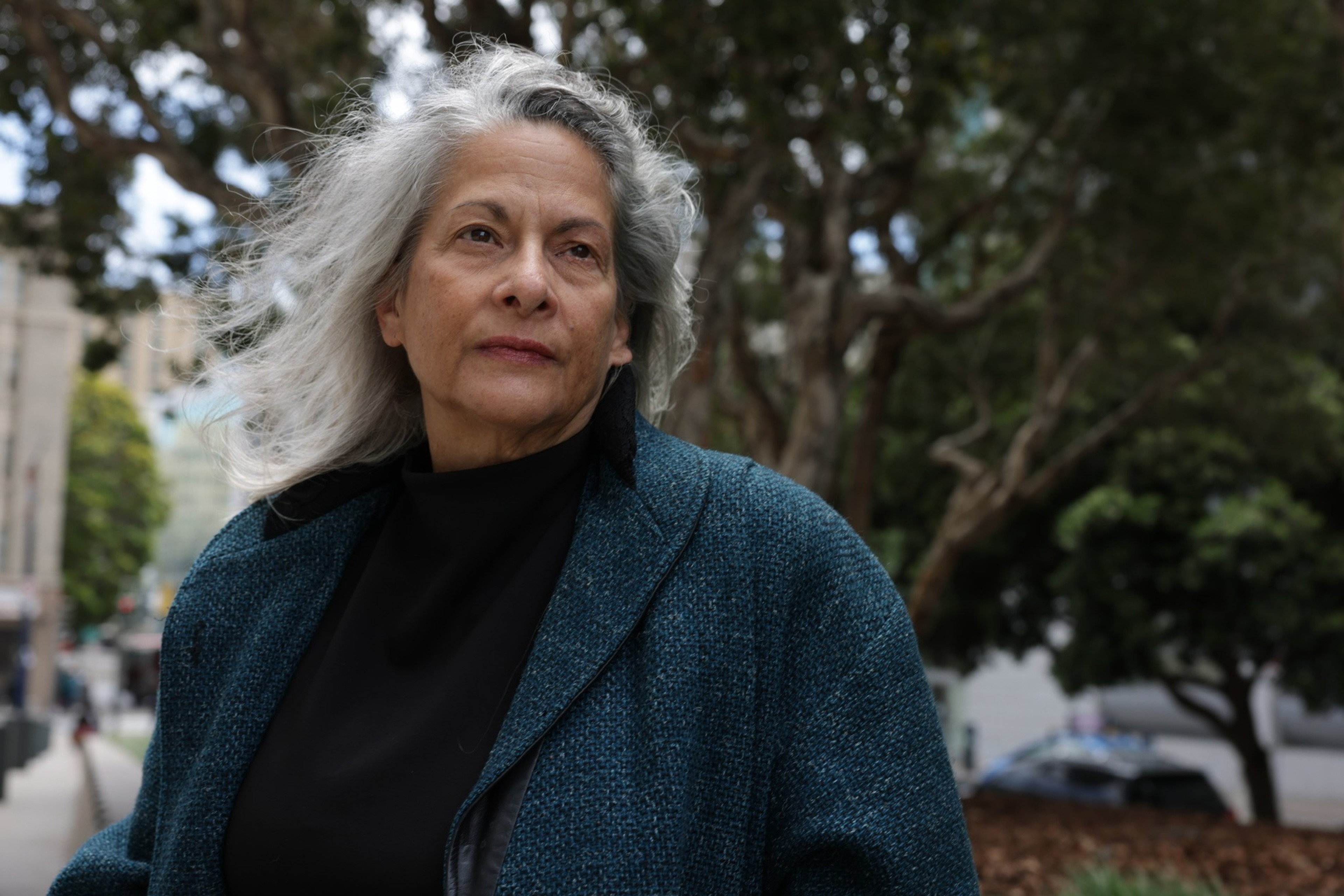 A woman with long gray hair, wearing a teal coat and black top, looks thoughtful while standing outdoors near trees with an urban backdrop.