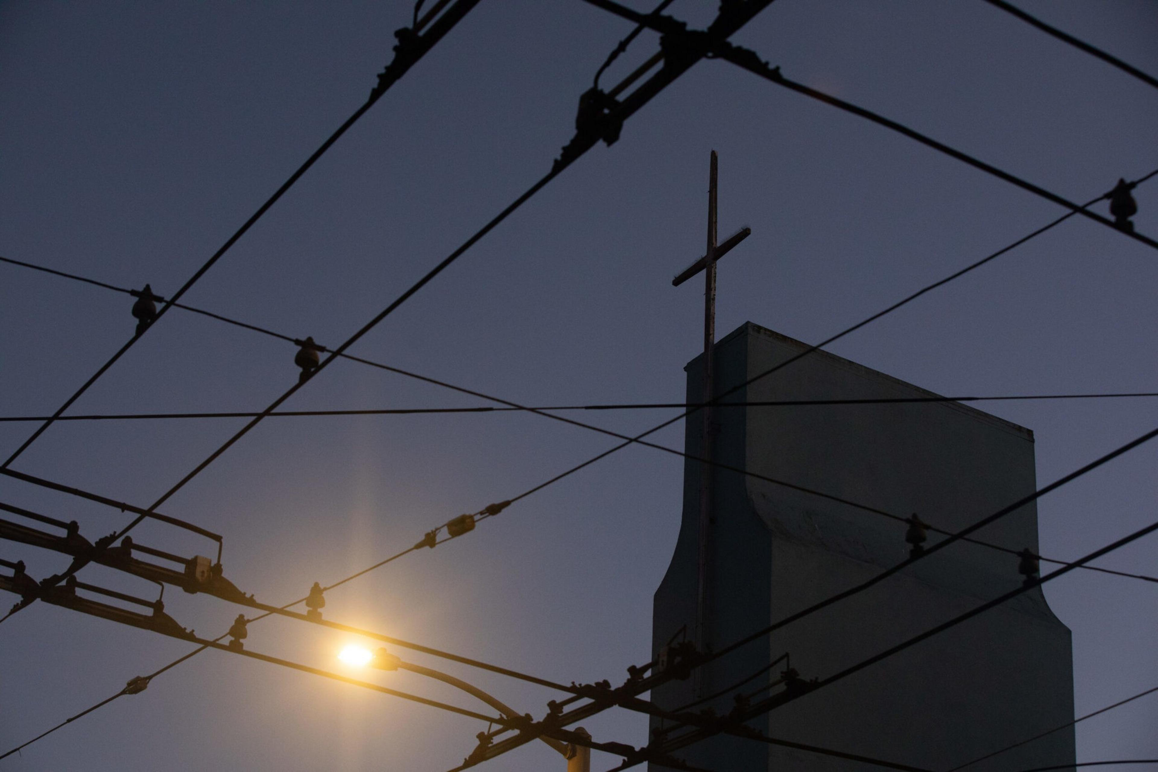 A church steeple with a cross is silhouetted against a dark sky, with numerous overhead utility wires crisscrossing in front.