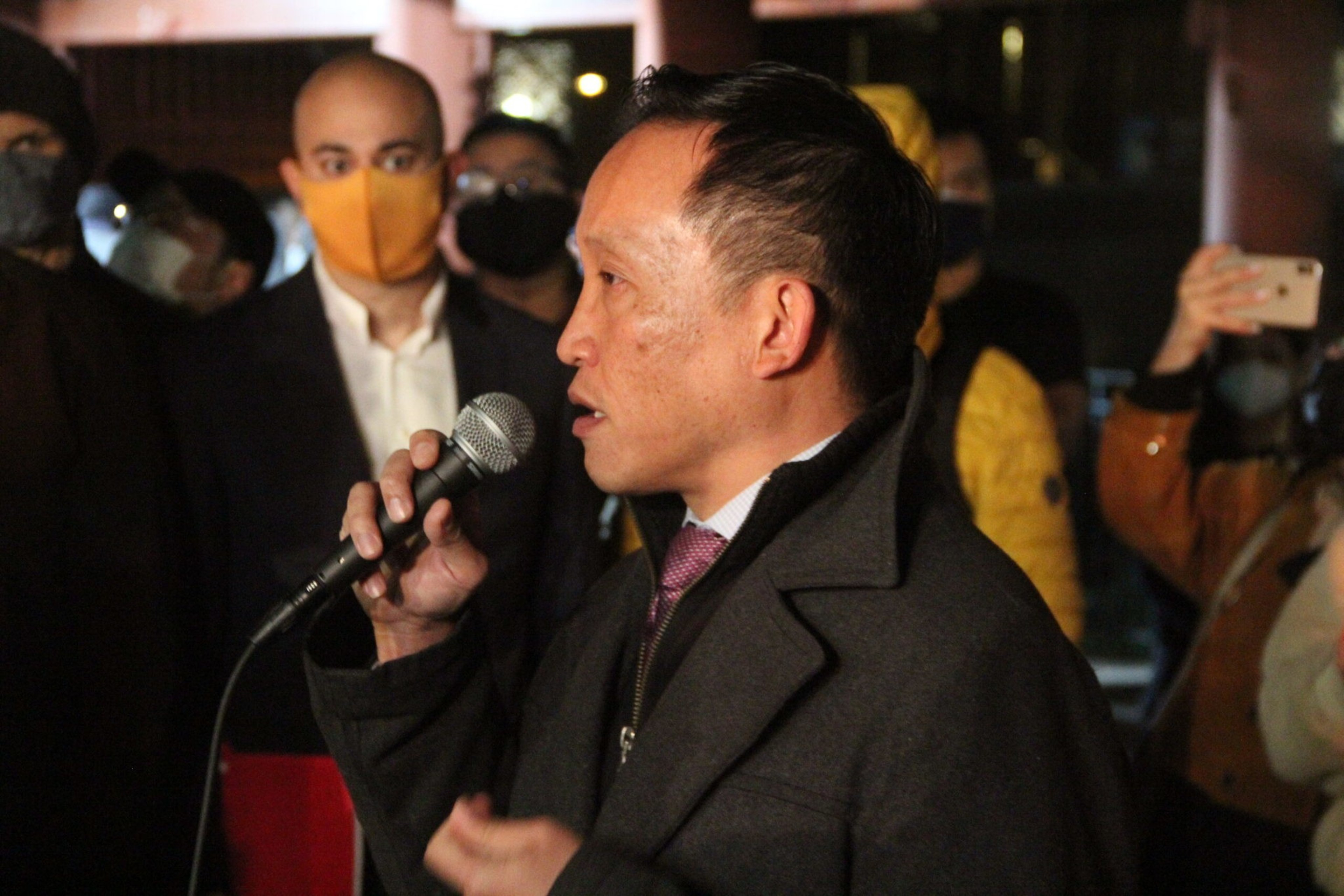 A man in a dark coat speaks into a microphone while several masked people stand behind him, one recording with a smartphone.