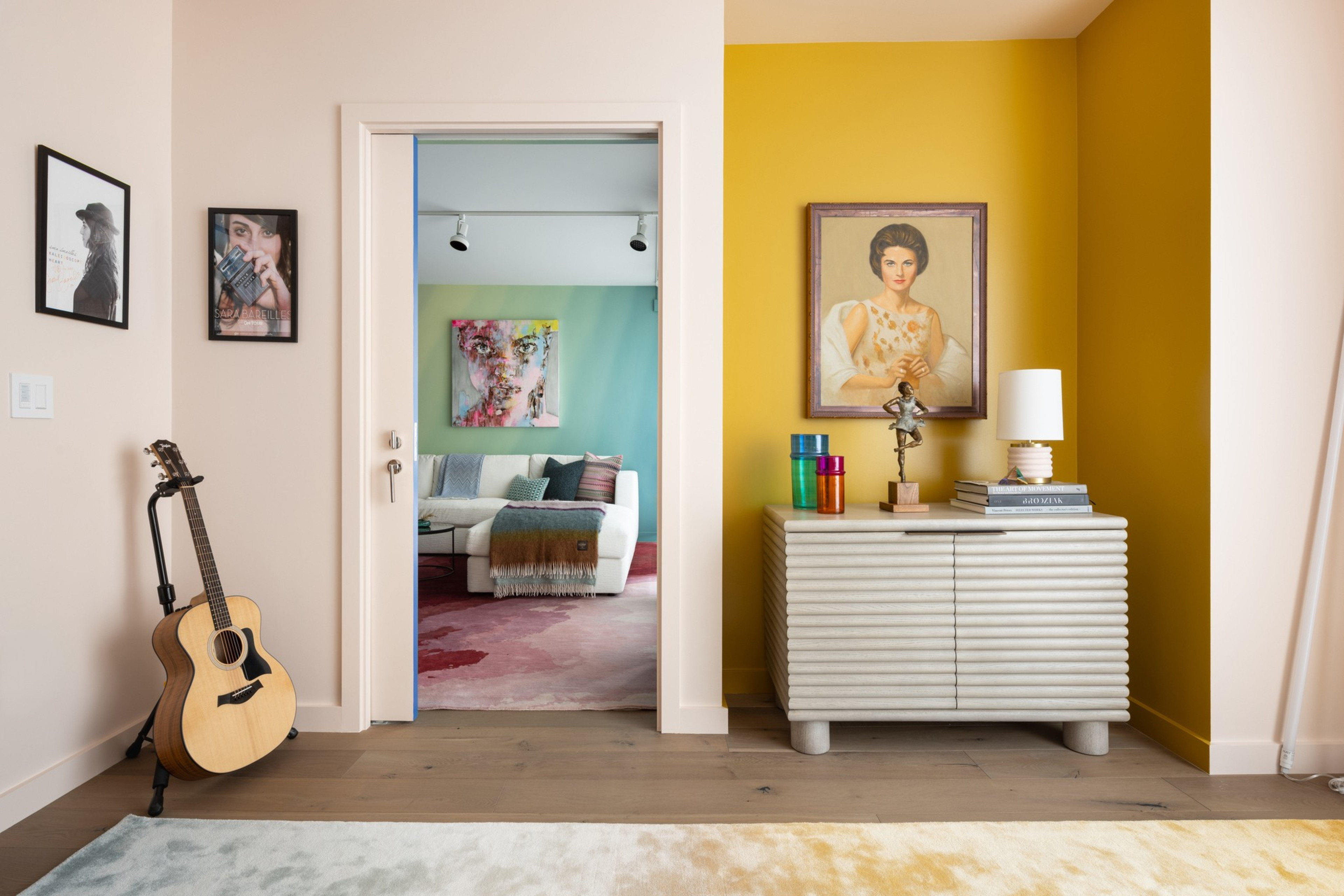 An acoustic guitar on a stand sits by a beige wall with two framed pictures, while a yellow wall holds a white cabinet topped with colorful glasses, a statue, books, lamp, and a portrait.