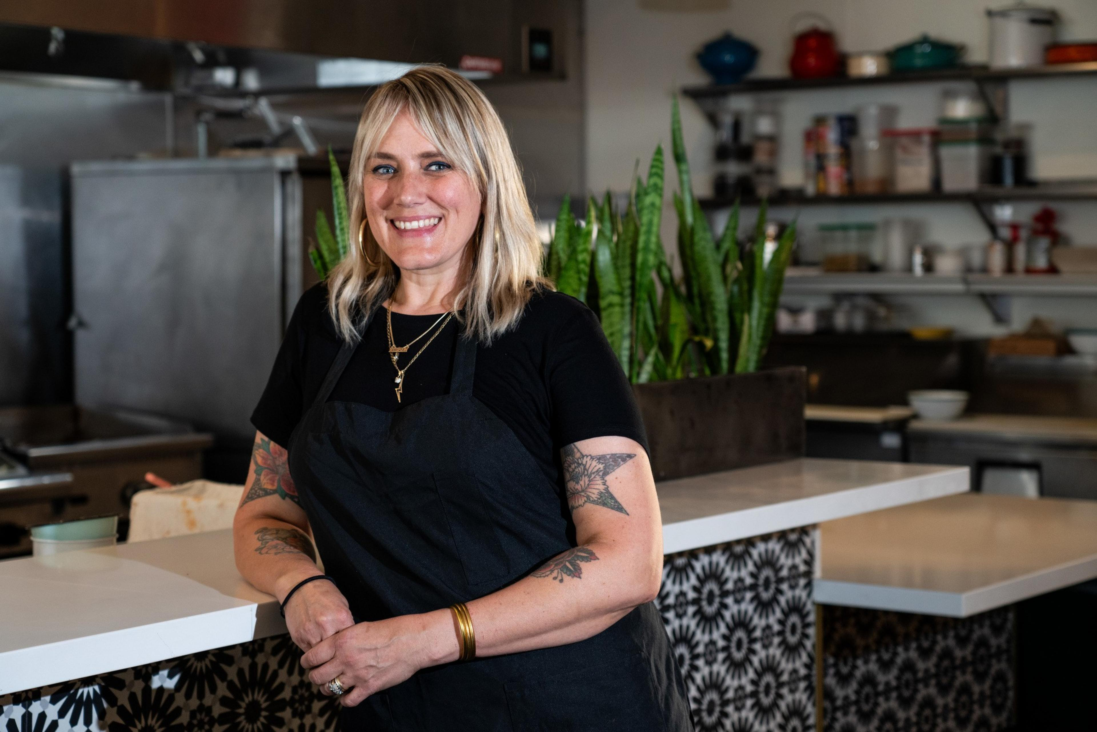A woman with blonde hair, tattoos, and a black apron stands smiling in a kitchen with shelves and plants behind her.