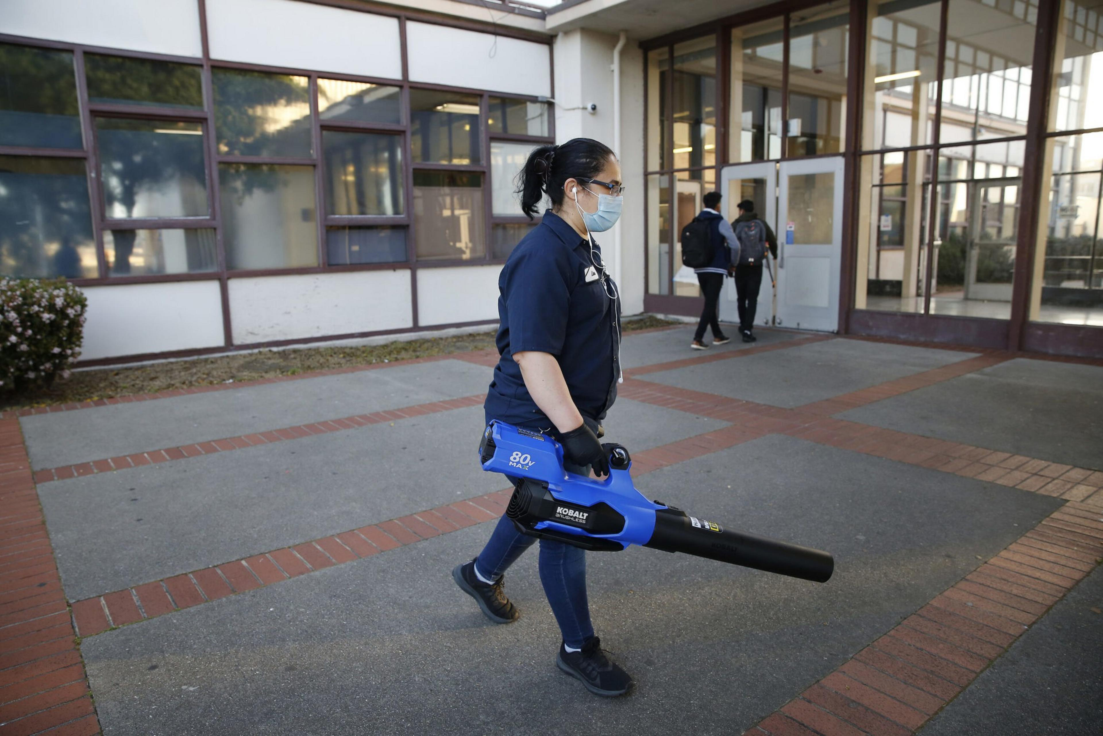 A person wearing a mask and gloves carries a blue leaf blower outside a building while two people walk into the entrance in the background.