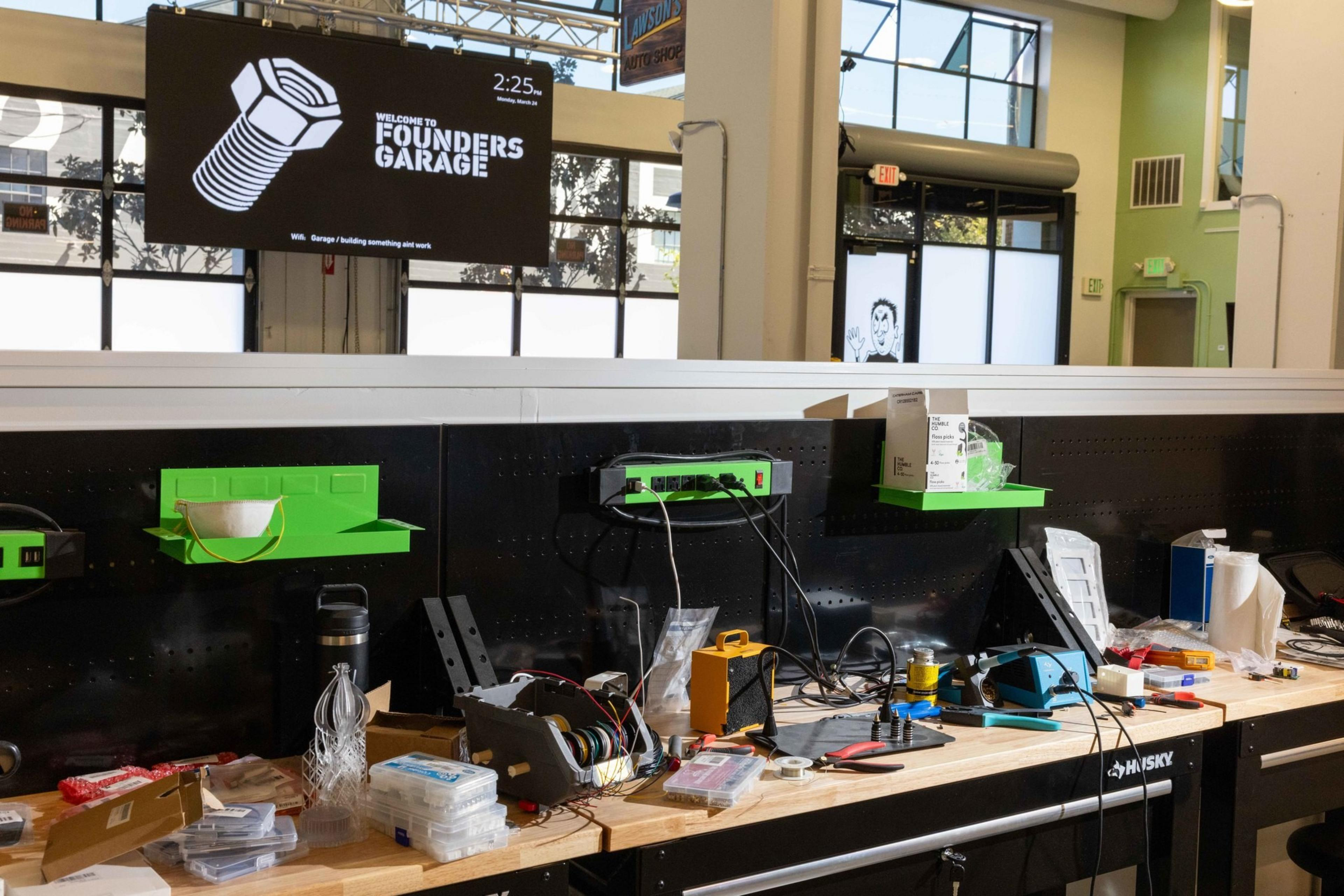The image shows a cluttered workshop with tools, wires, and electronic equipment on a wooden bench. A &quot;Founders Garage&quot; sign hangs above, and sunlight streams in through large windows.