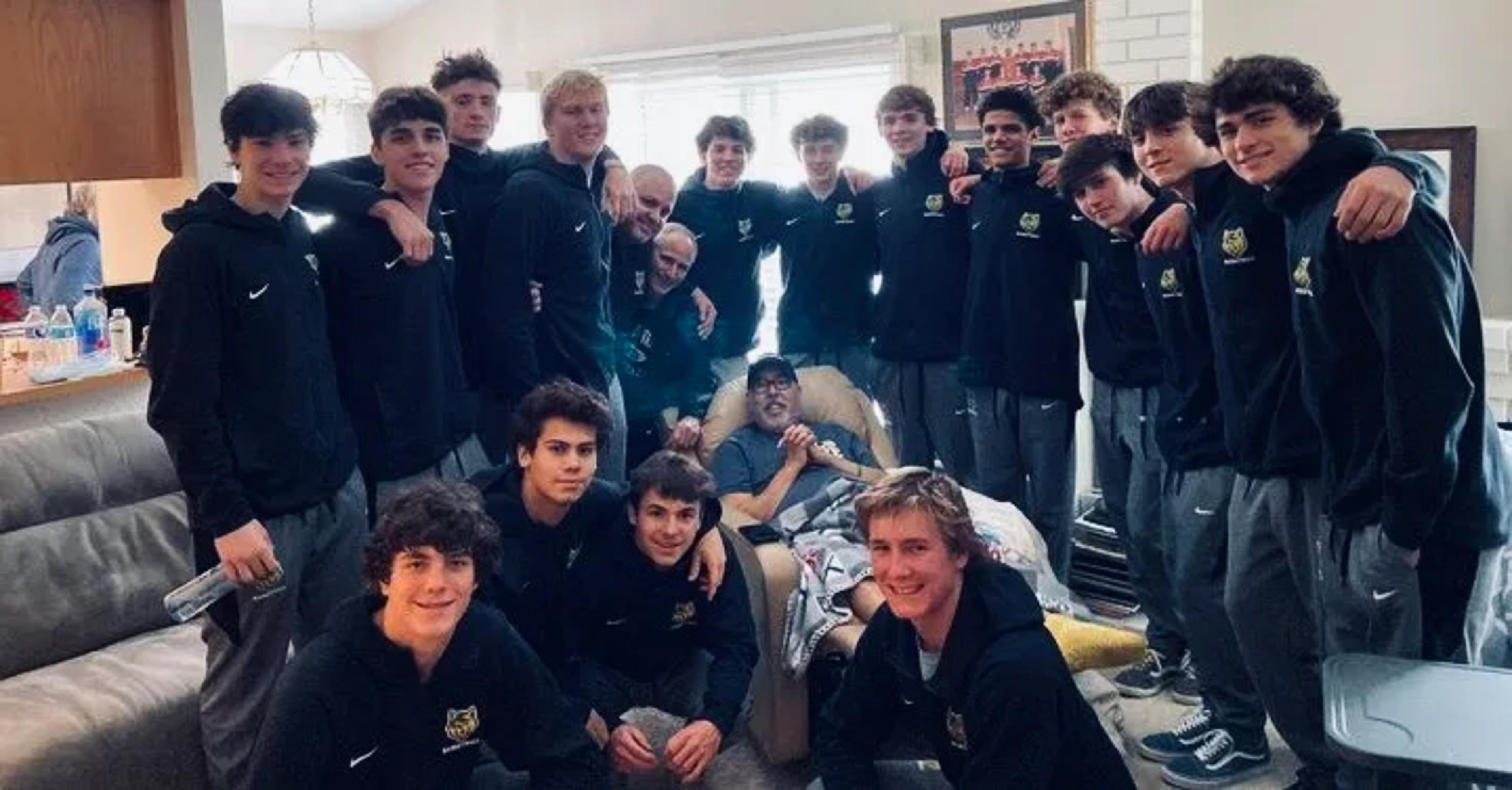 A group of young men in matching black hoodies gather around two men seated on a chair and recliner in a cozy living room.
