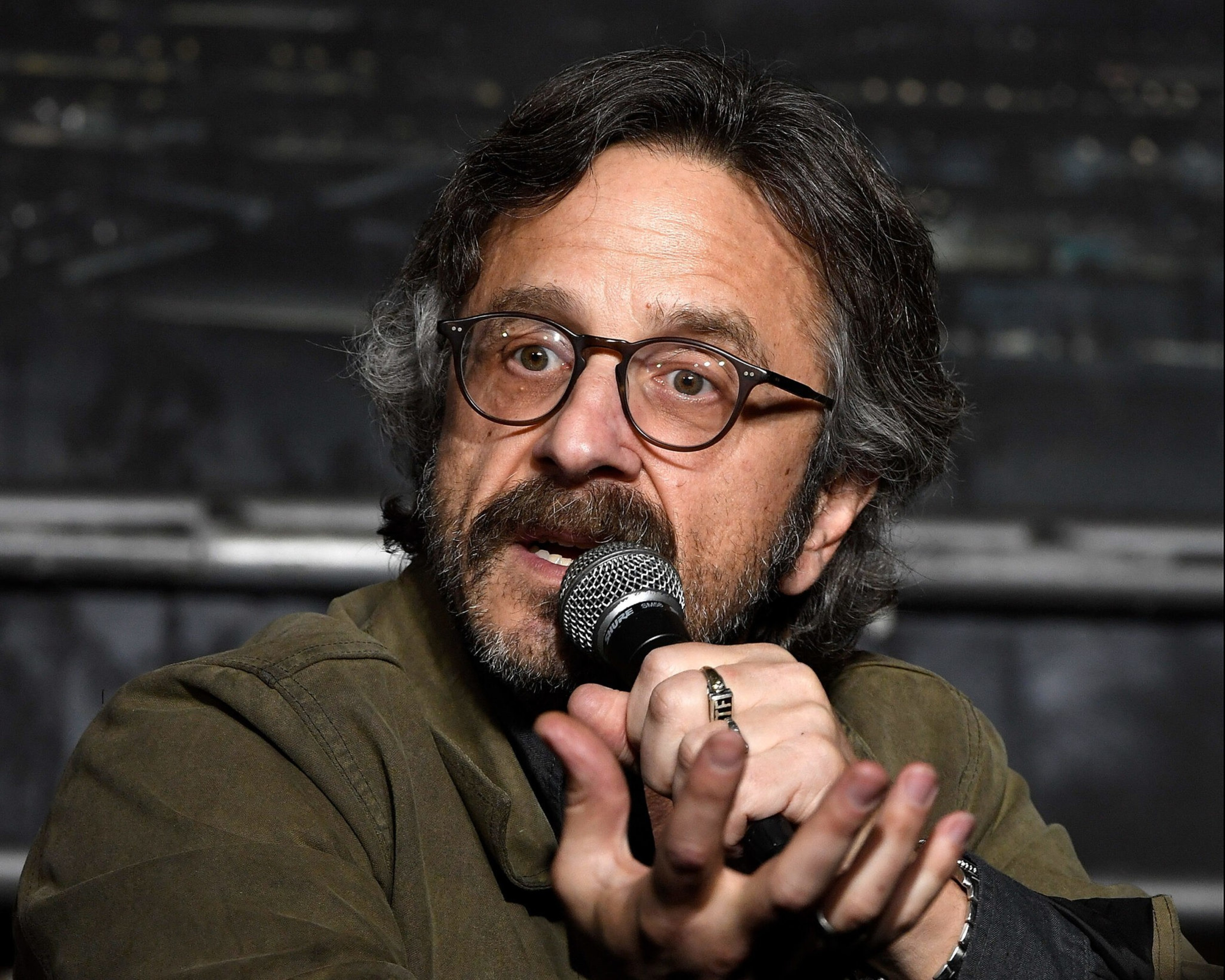 A man with glasses, curly hair, and a beard speaks into a microphone, gesturing with one hand while wearing a green jacket.