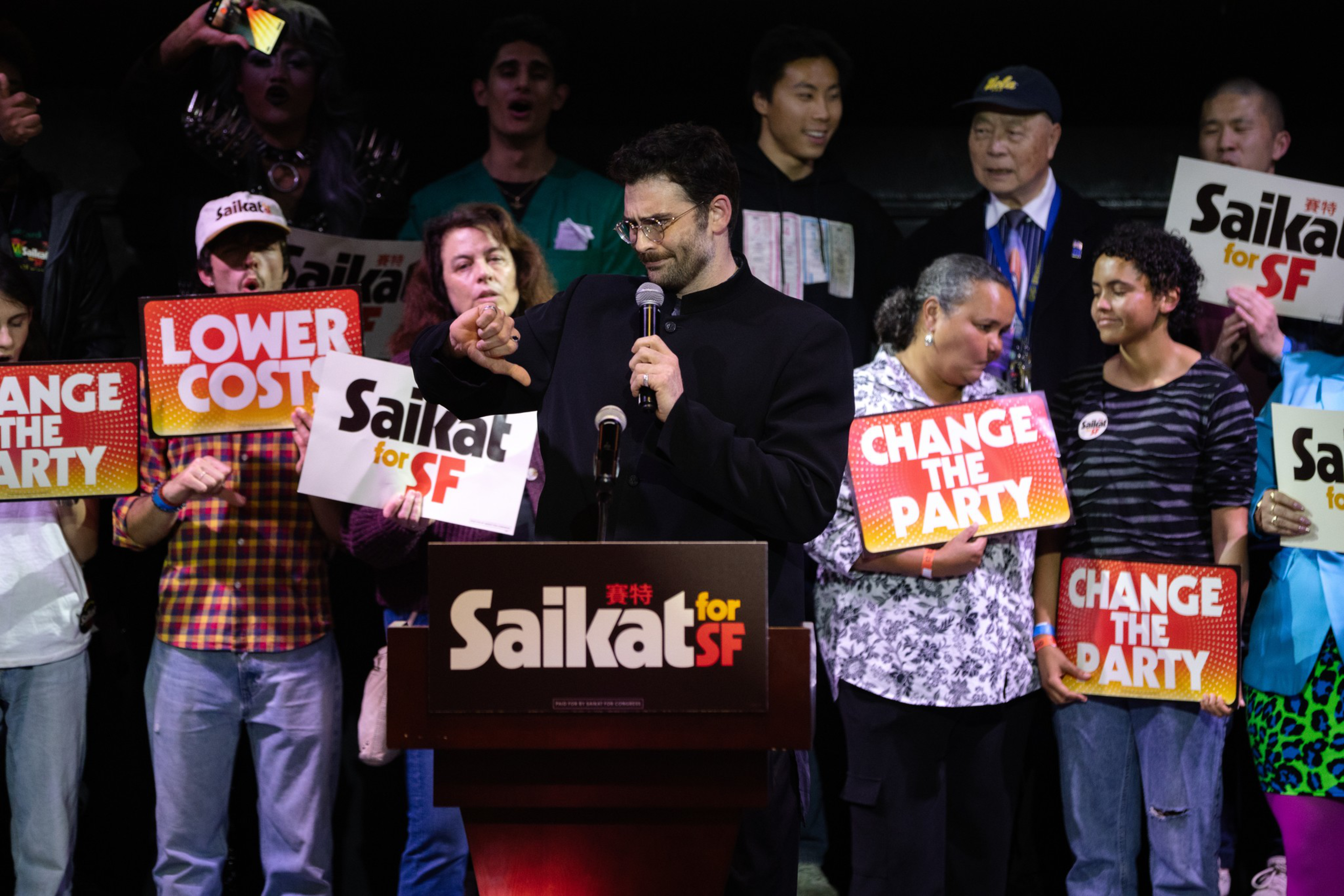 A man speaks at a podium labeled “Saikat for SF,” surrounded by diverse supporters holding signs saying “CHANGE THE PARTY,” “LOWER COST,” and “Saikat for SF.”