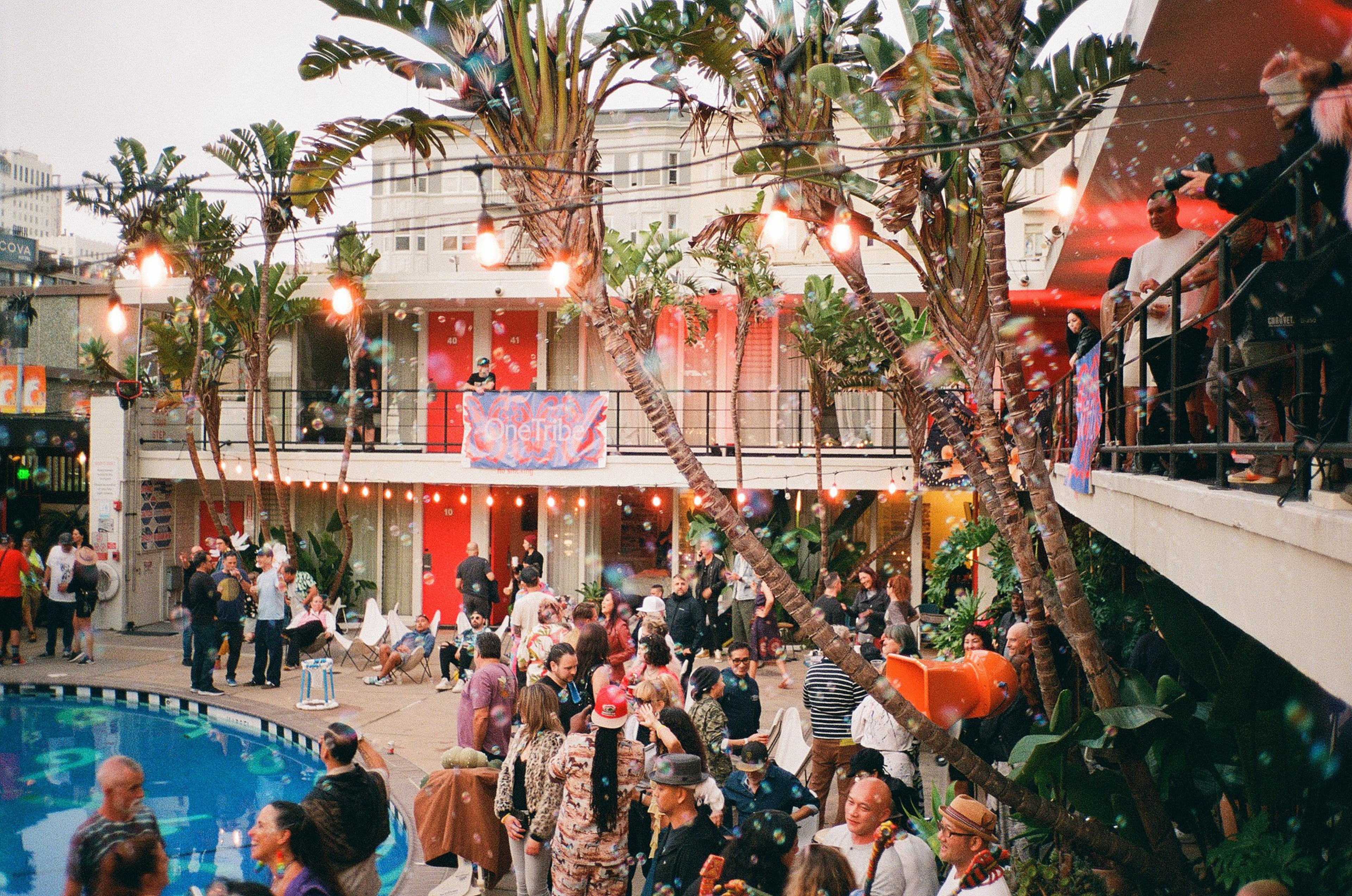 A lively outdoor party with many people mingling around a pool, surrounded by palm trees, string lights, and a two-story building with red doors.