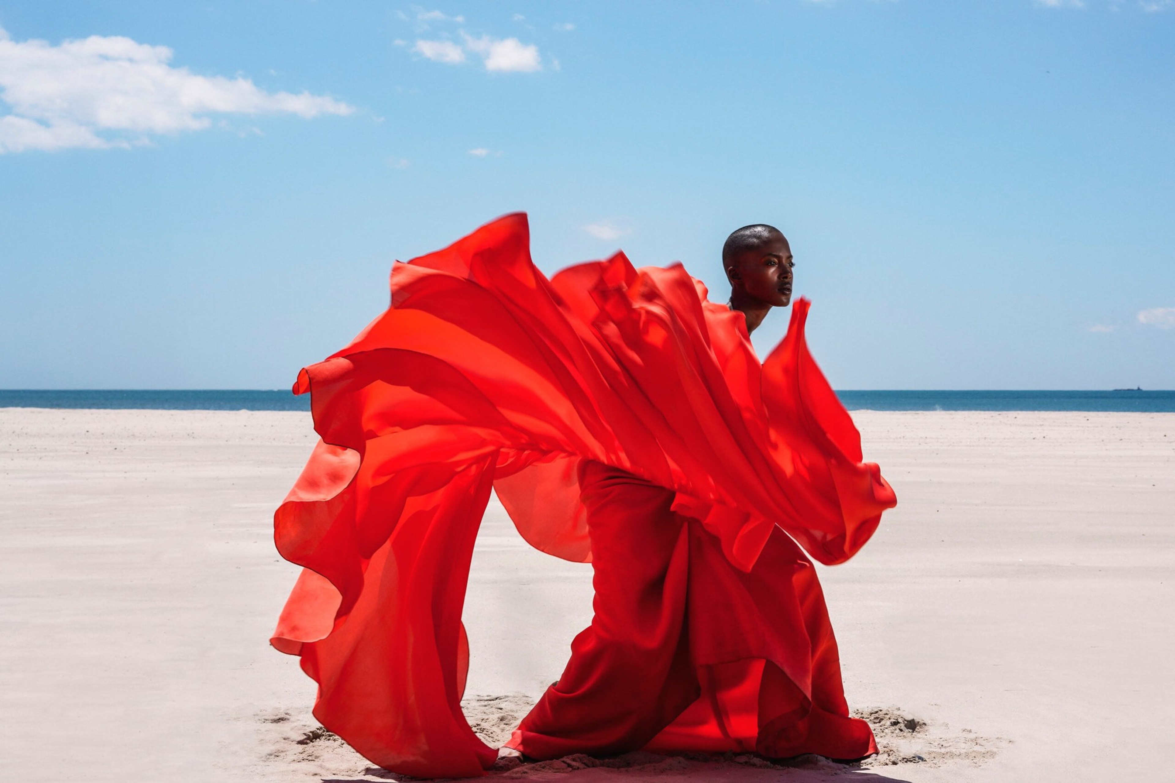 A person stands on a vast sandy beach wearing a flowing bright red dress that billows dramatically against a clear blue sky.