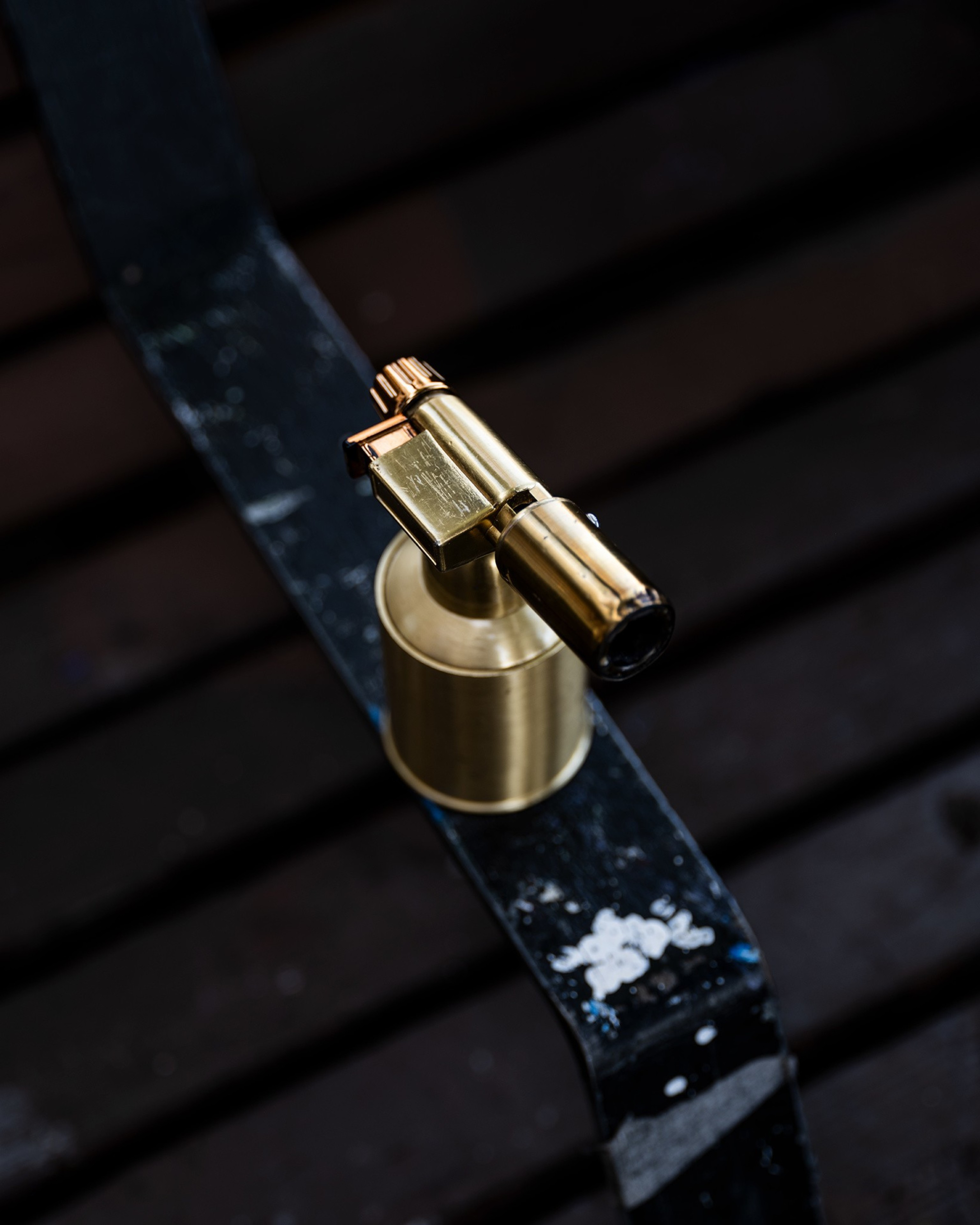 A small, metallic blowtorch with a golden finish sits on a dark, textured surface with visible paint scratches.