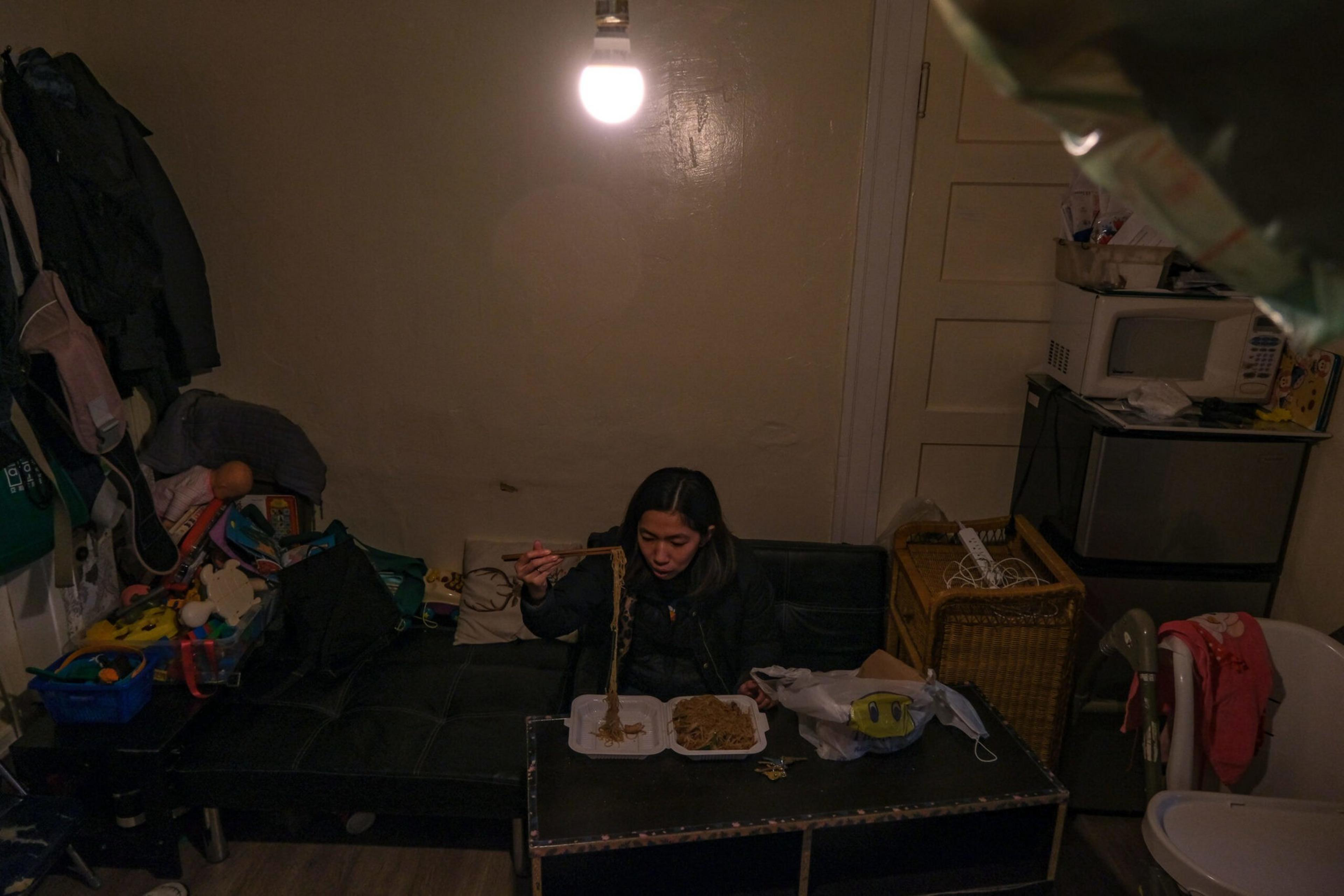 A person sits on a dark couch under a single hanging light, eating noodles with chopsticks in a cluttered, dimly lit room.
