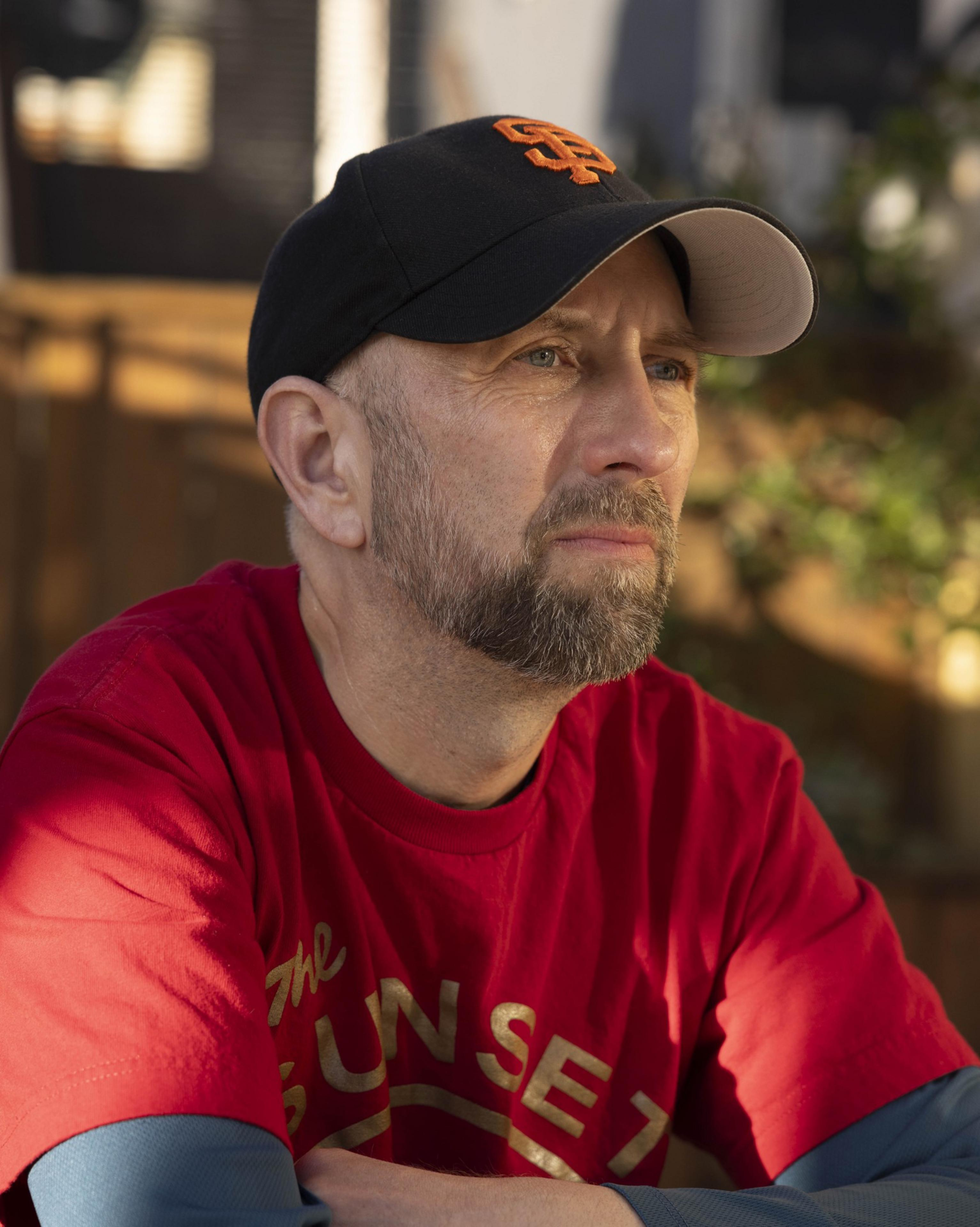 A bearded man in a black baseball cap with orange lettering gazes thoughtfully into the distance, wearing a red &quot;SUNSET&quot; T-shirt in a sunlit outdoor area.