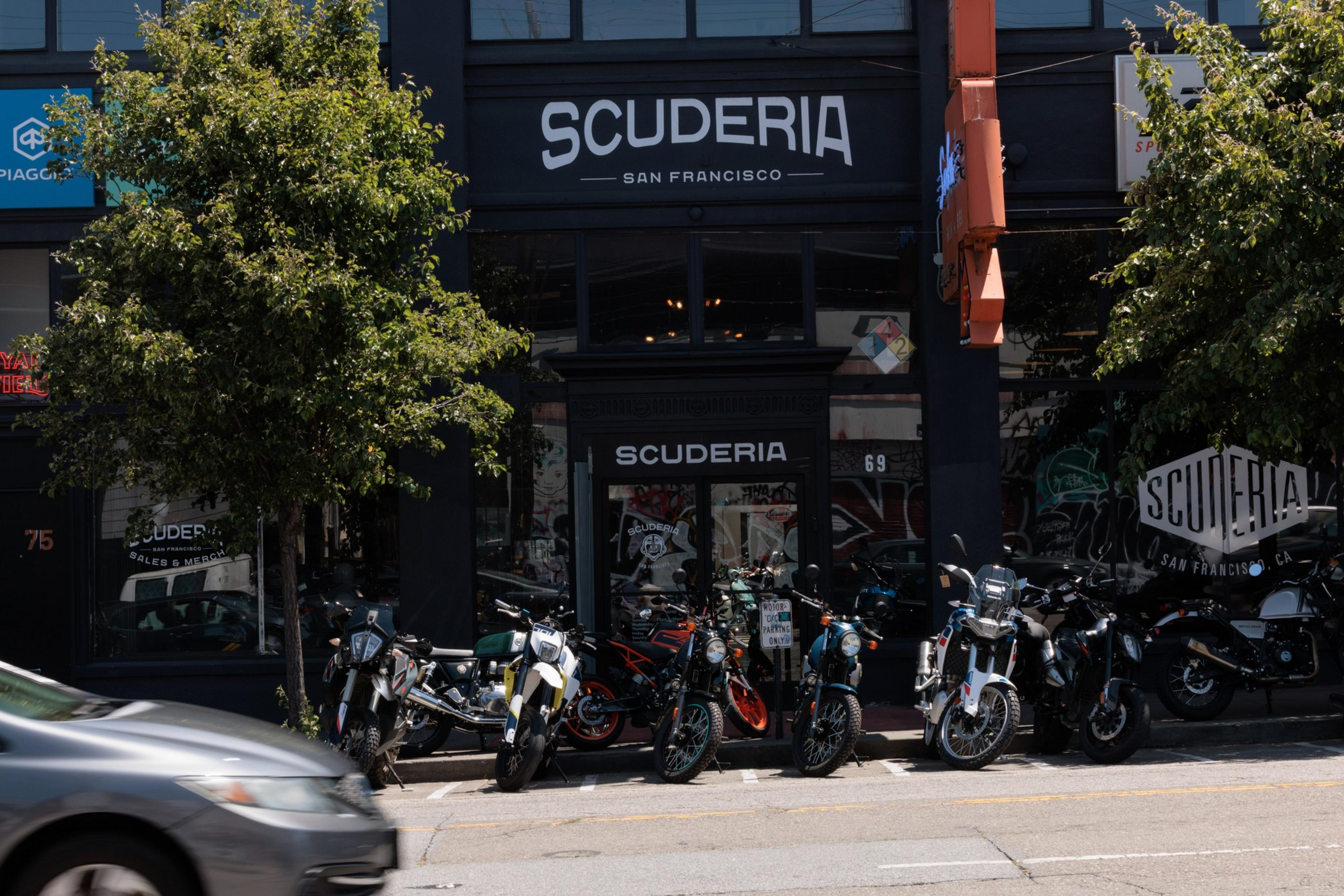 A motorcycle shop named "SCUDERIA San Francisco" with several motorcycles parked in front on a sunny day, flanked by two leafy trees.
