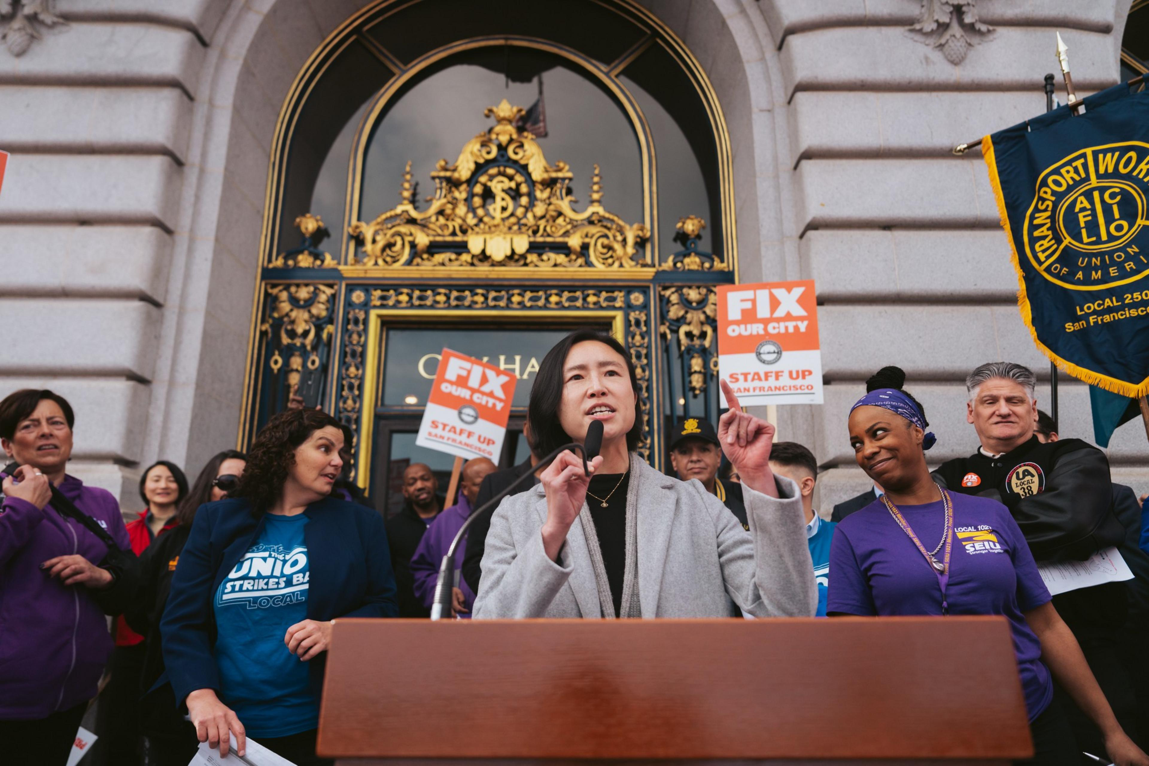 A person speaks at a podium before a group, some with union signs, outside an ornate building.