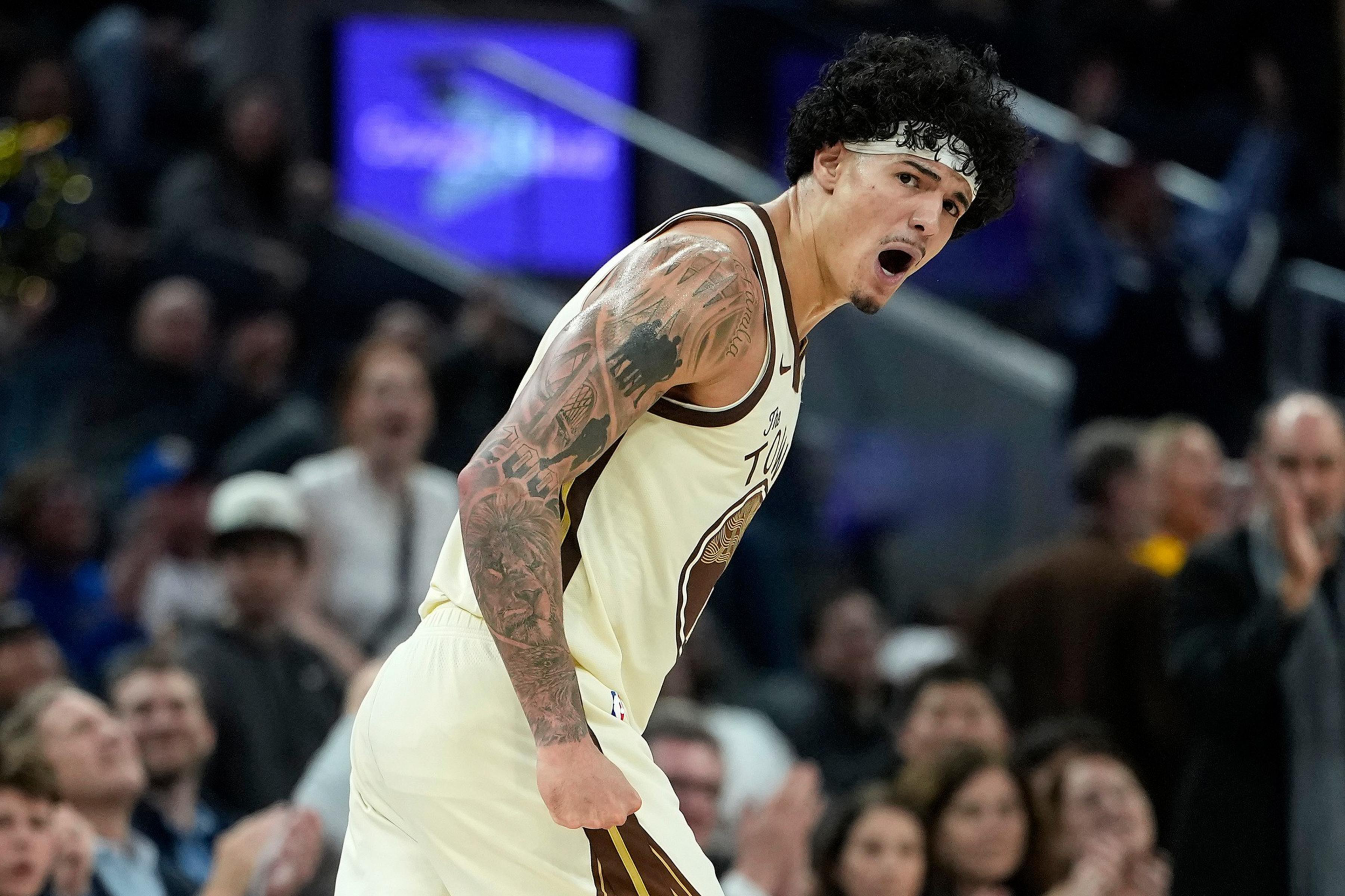A basketball player with tattoos and a headband is shouting passionately during a game, with a crowd blurred in the background.
