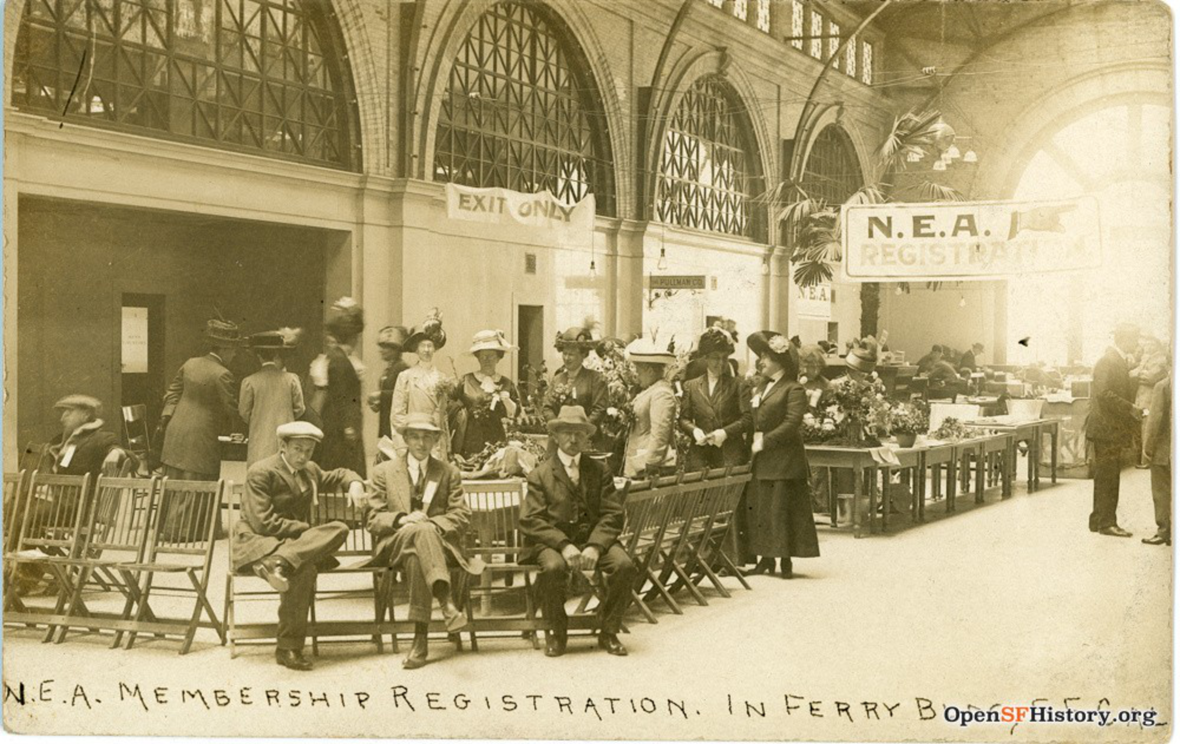 People in early 20th-century clothing wait or register at a long table under signs reading "N.E.A. Membership Registration" in a large, arched ferry building.