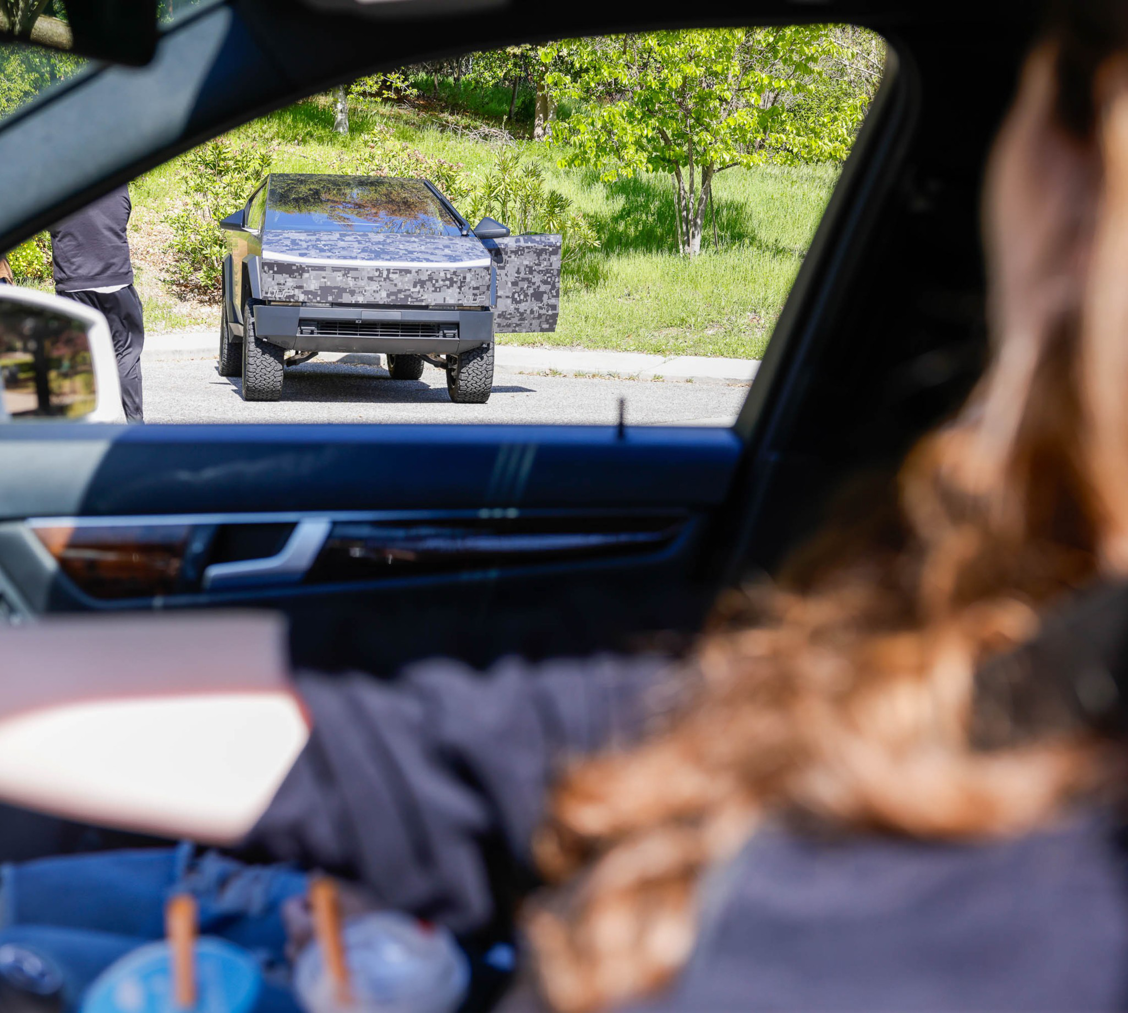 A person in a car is viewing another person opening the door of a camouflaged angular vehicle.