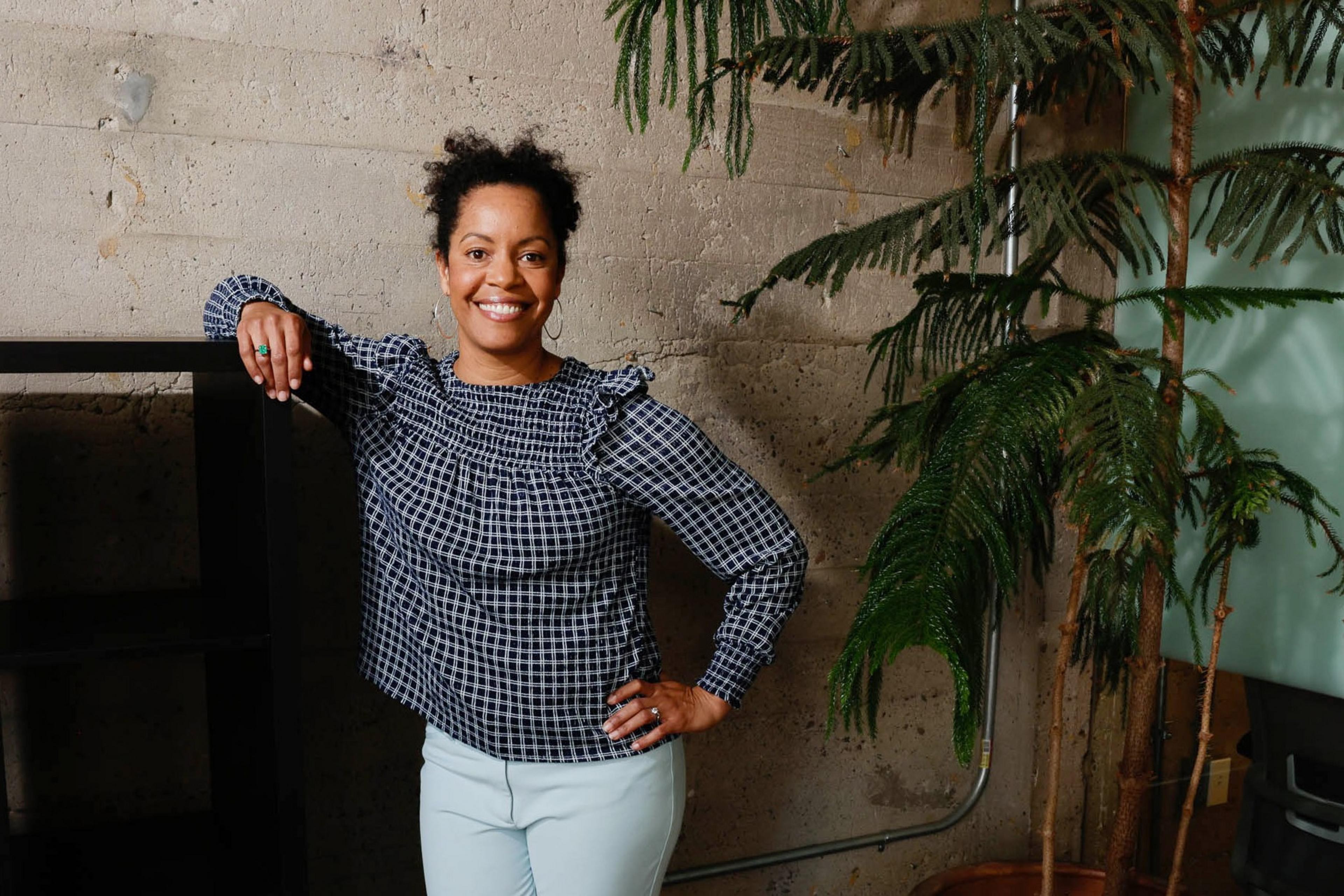 A smiling woman with a checkered top and light pants stands by a bookshelf and a plant.