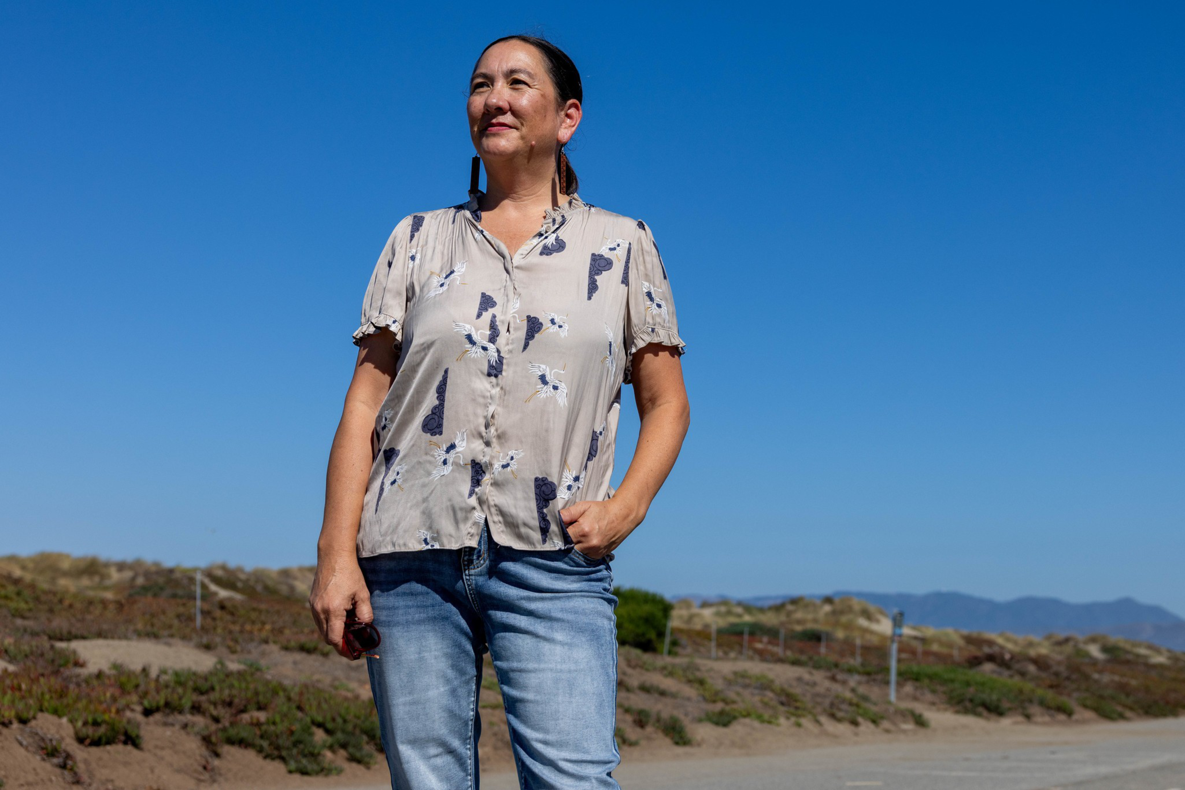 A woman with dark hair tied back stands outdoors against a clear blue sky, wearing a patterned blouse and jeans, with one hand in her pocket.